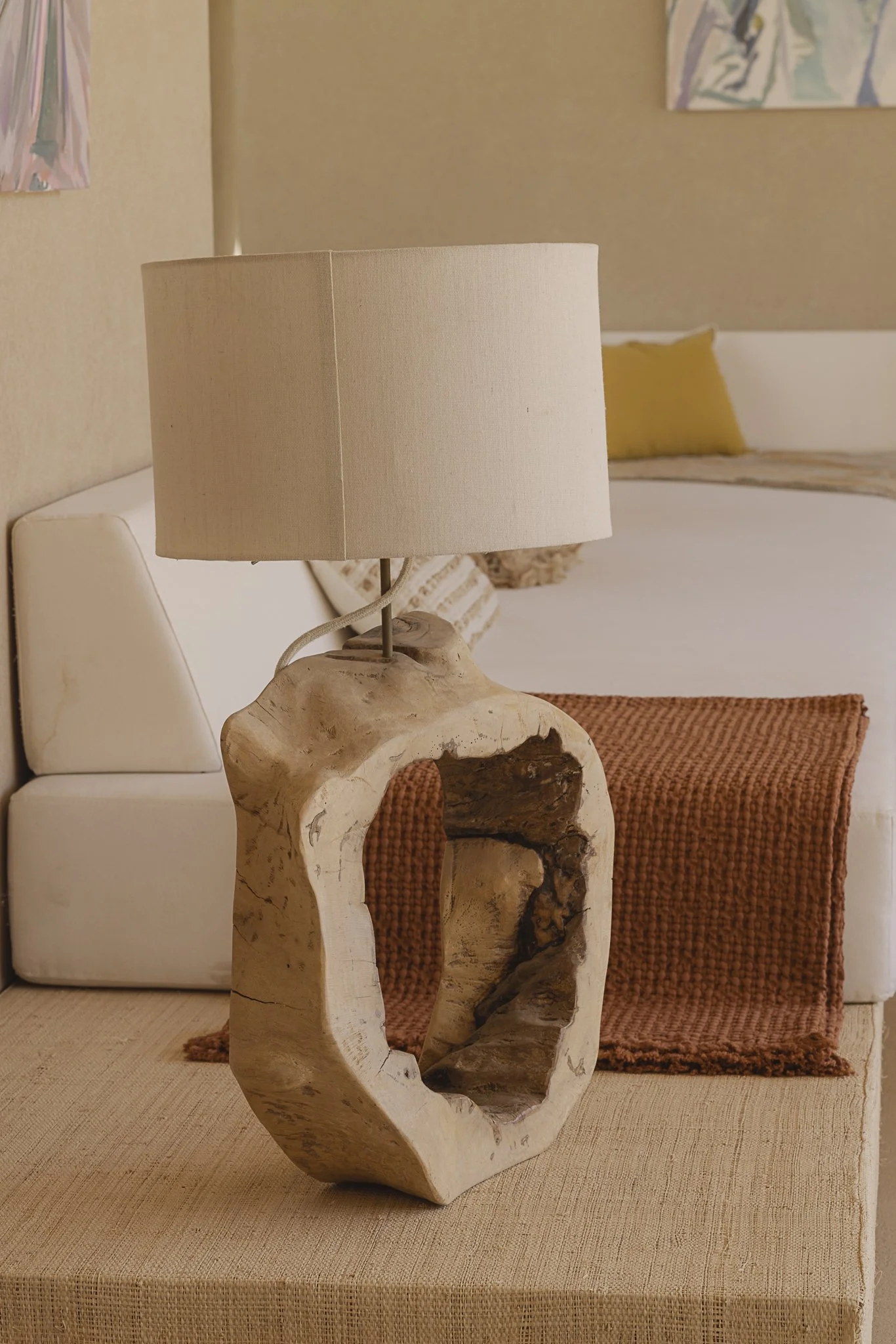 A beige table lamp on a beige table, with a large hollow wooden base and a cream lampshade, placed in front of a white couch with a rust-colored throw pillow and a yellow pillow, in a cozy living room with abstract art on the wall.