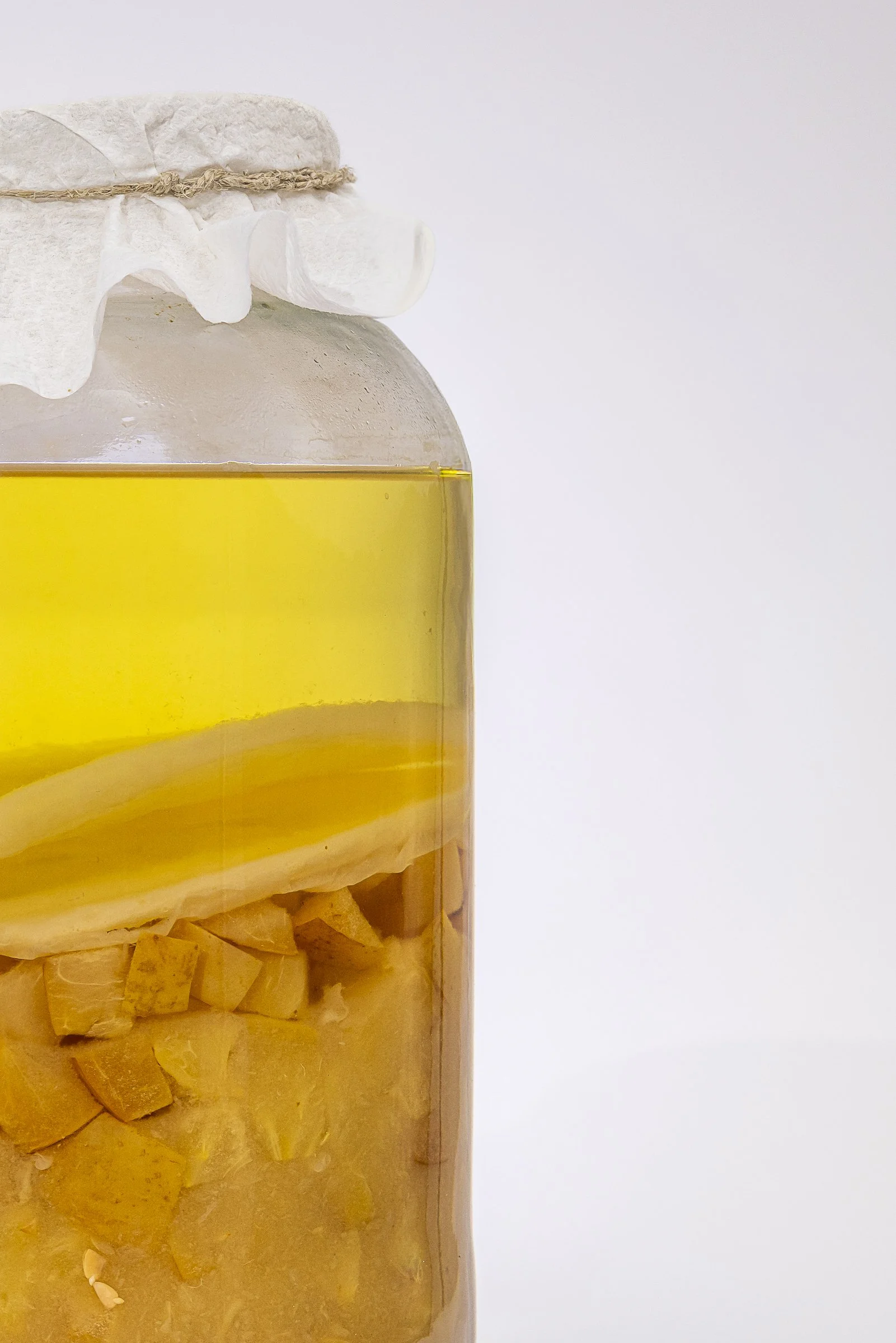 A glass jar filled with yellow liquid and chopped yellow fruit pieces, topped with a paper cover secured with a string, against a plain white background.