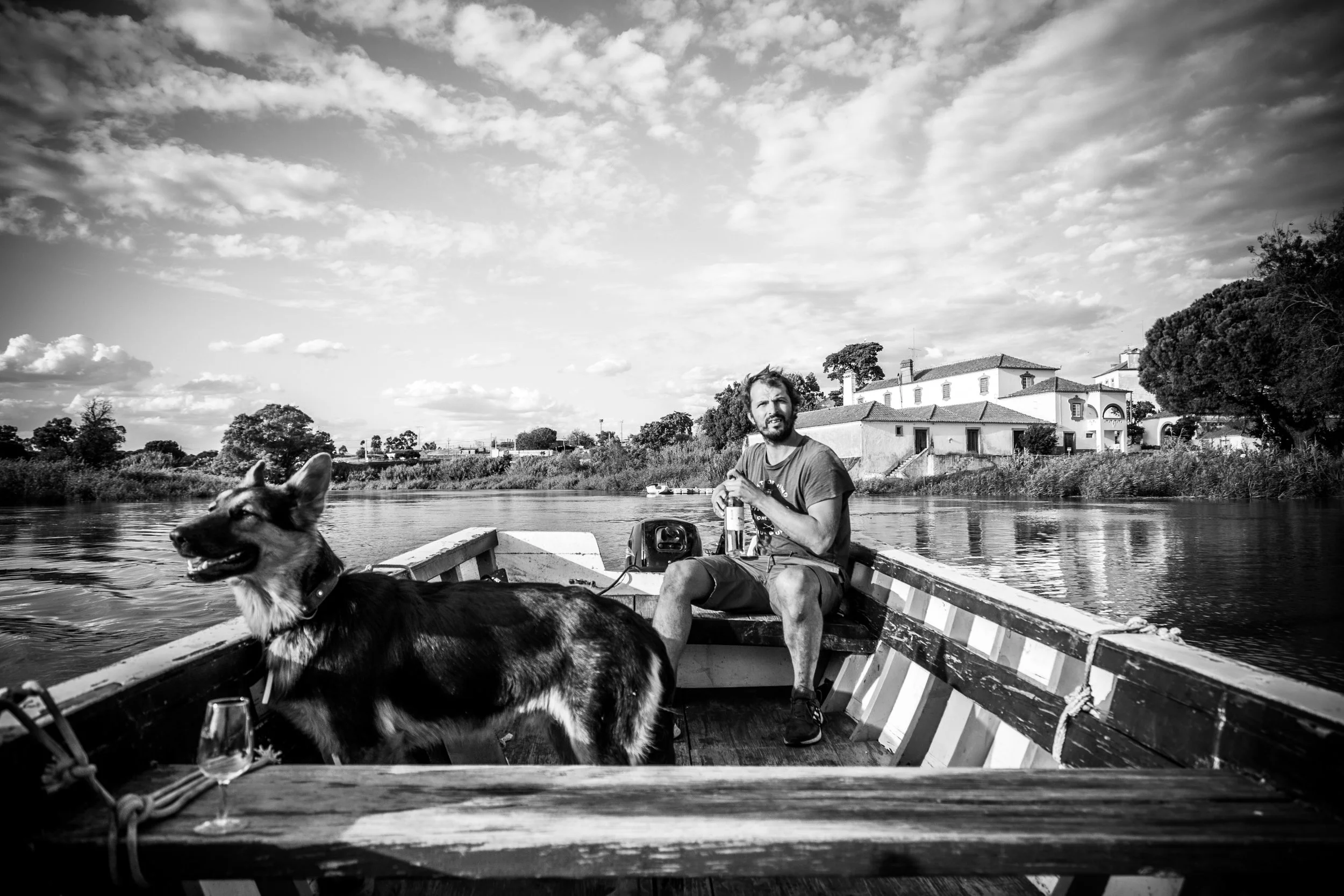 A man and a dog sitting in a small boat on a river with houses and trees in the background, under a partly cloudy sky.