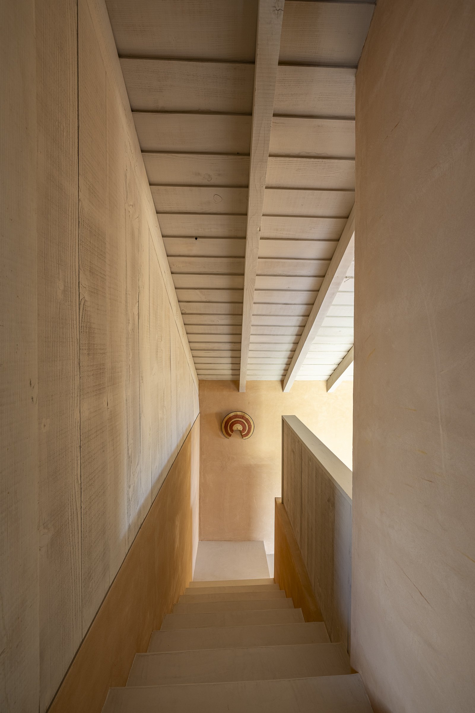 Wooden staircase viewed from above with warm-toned walls and ceiling, featuring a decorative round wall hanging at the bottom.