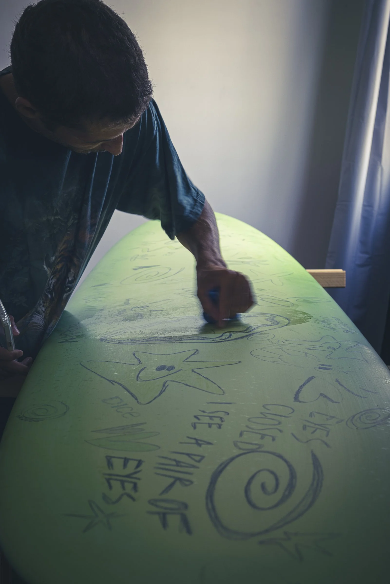 Person drawing on a green surfboard with various doodles and words, near a window with blue curtains.