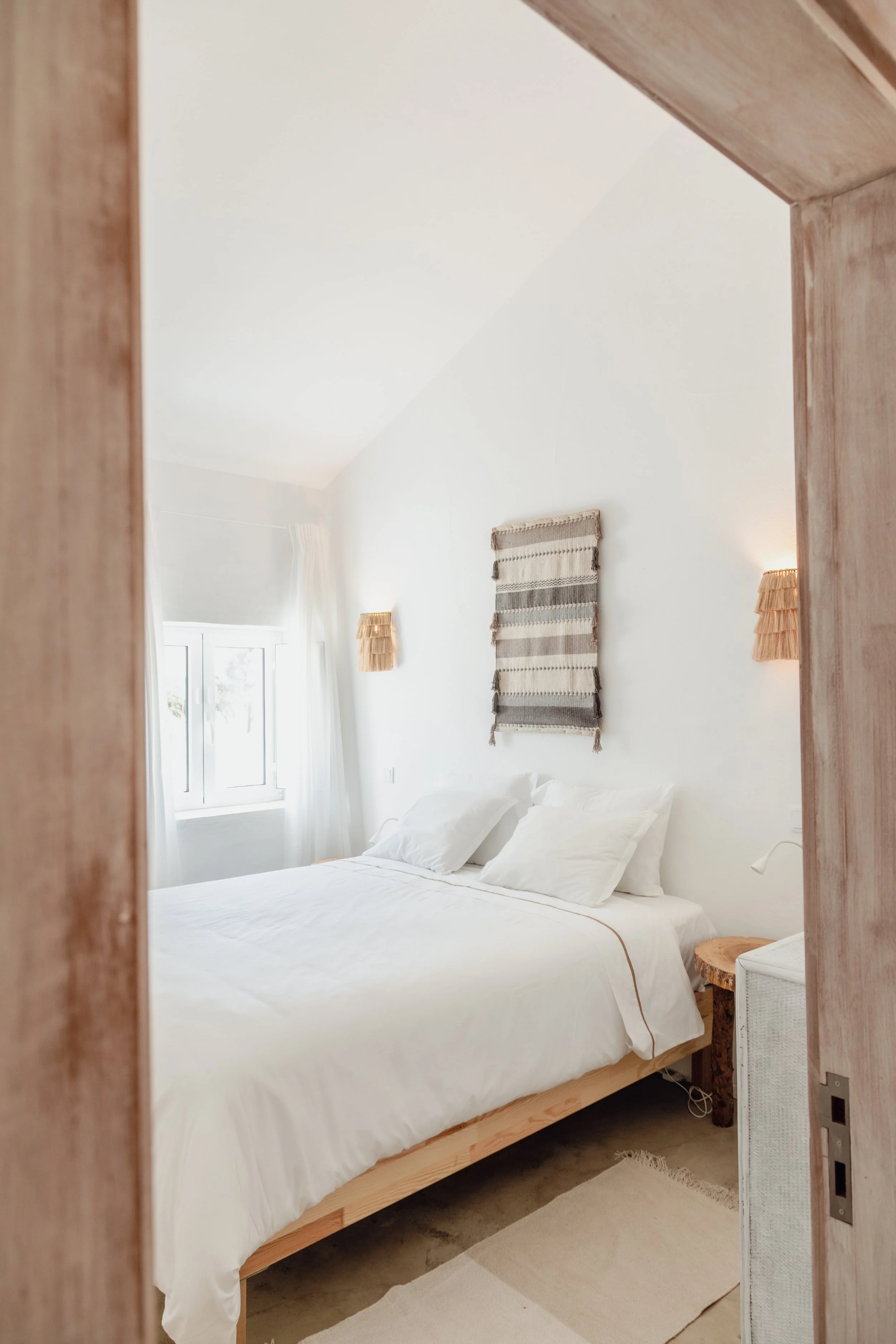 A cozy bedroom with white walls, a bed with white bedding and pillows, a window with white curtains, two wall-mounted lamps, a woven wall hanging, a wooden bedside table, and a beige rug.