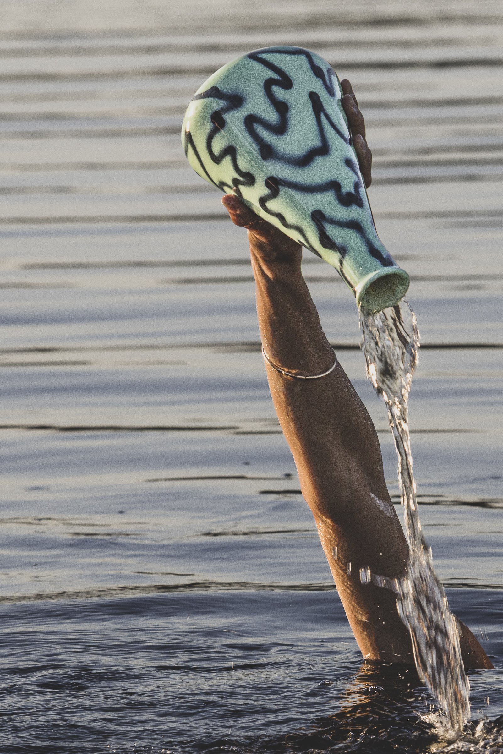 A person with dark skin is raising a blue and green patterned vase out of the water, with water pouring from it, in a body of water during sunset.