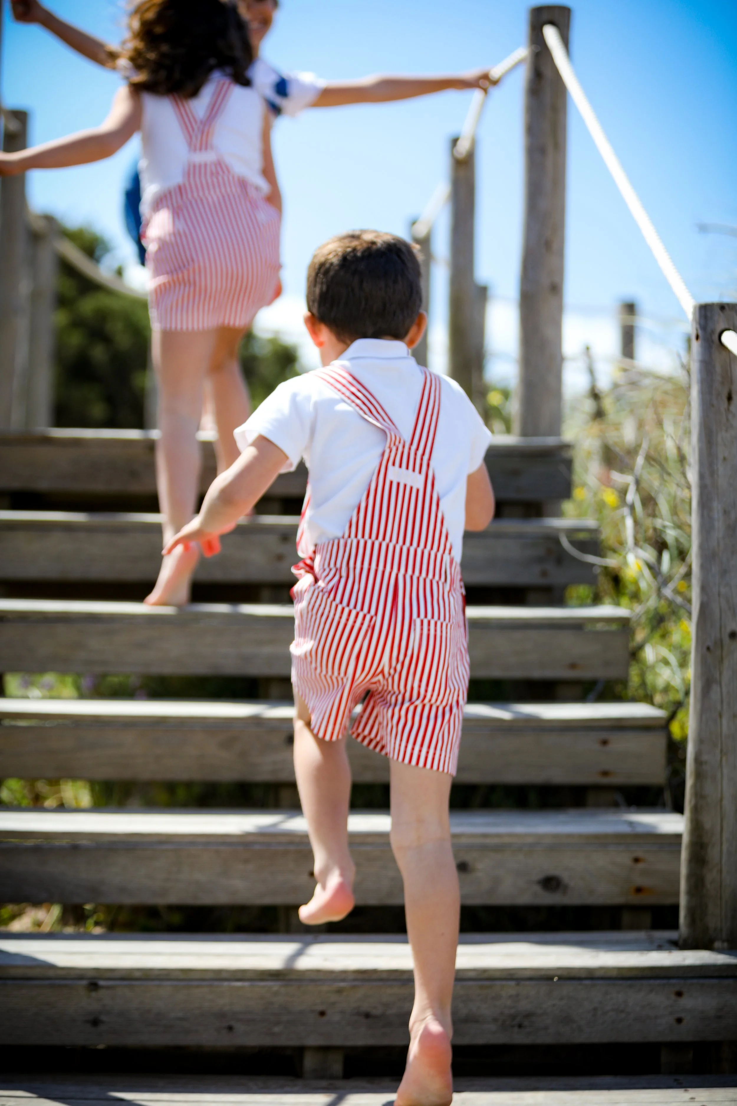 Children climbing outdoor wooden stairs; one boy with striped red and white overalls and another girl in similar attire at the top of the stairs with arms outstretched.