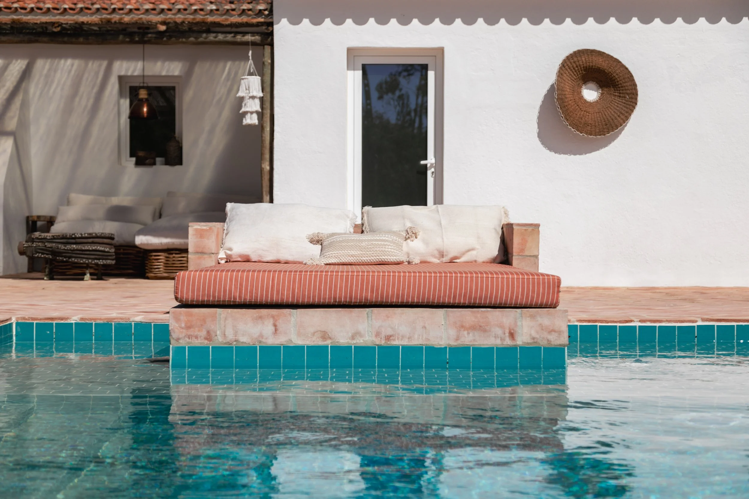 A poolside outdoor seating area with a cushioned lounge on the edge of a swimming pool, a white building with a door behind it, and decorative wall hangings.