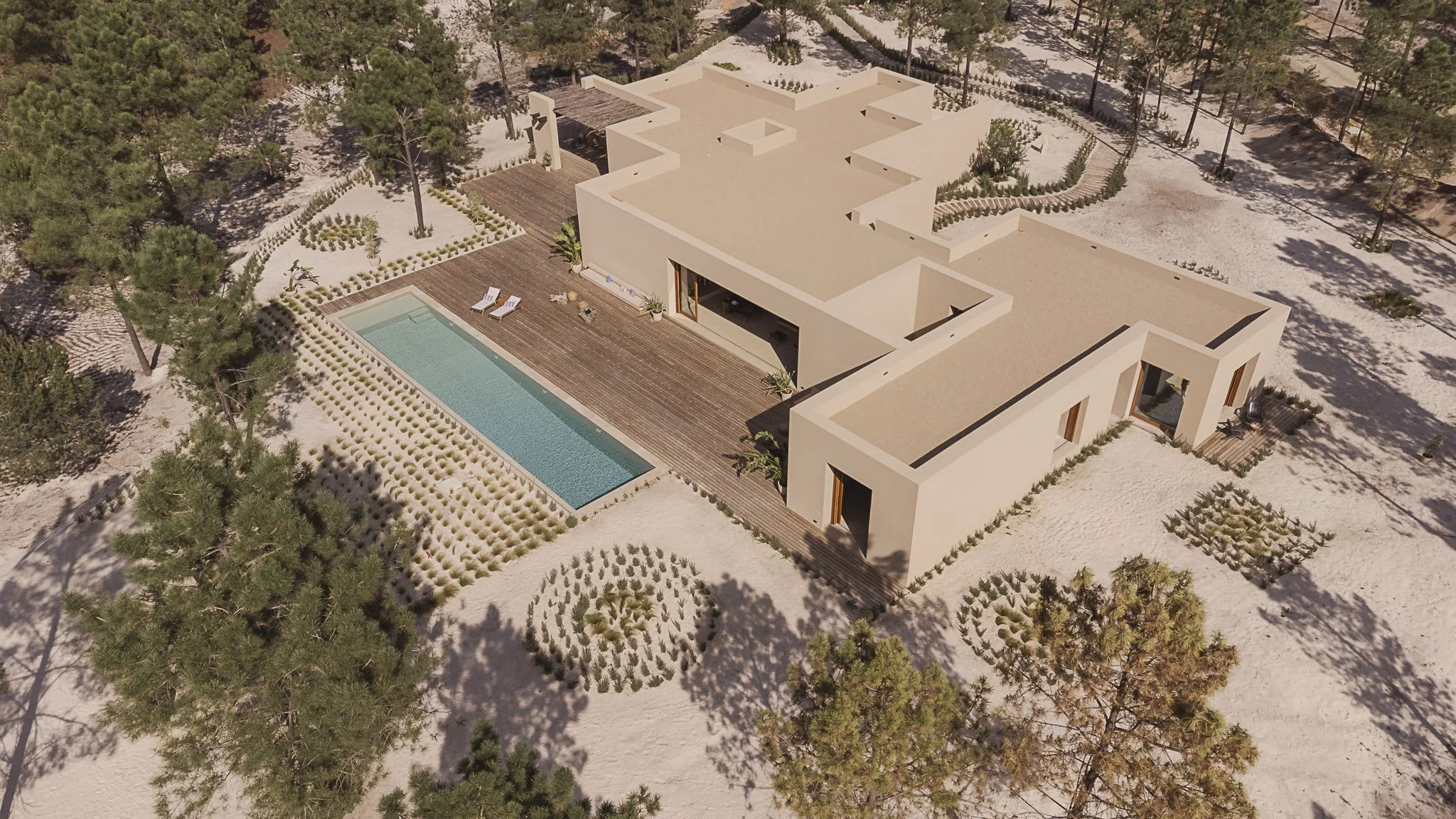A modern house with flat roofs, surrounded by sandy terrain and trees, featuring a swimming pool on a wooden deck with lounge chairs.
