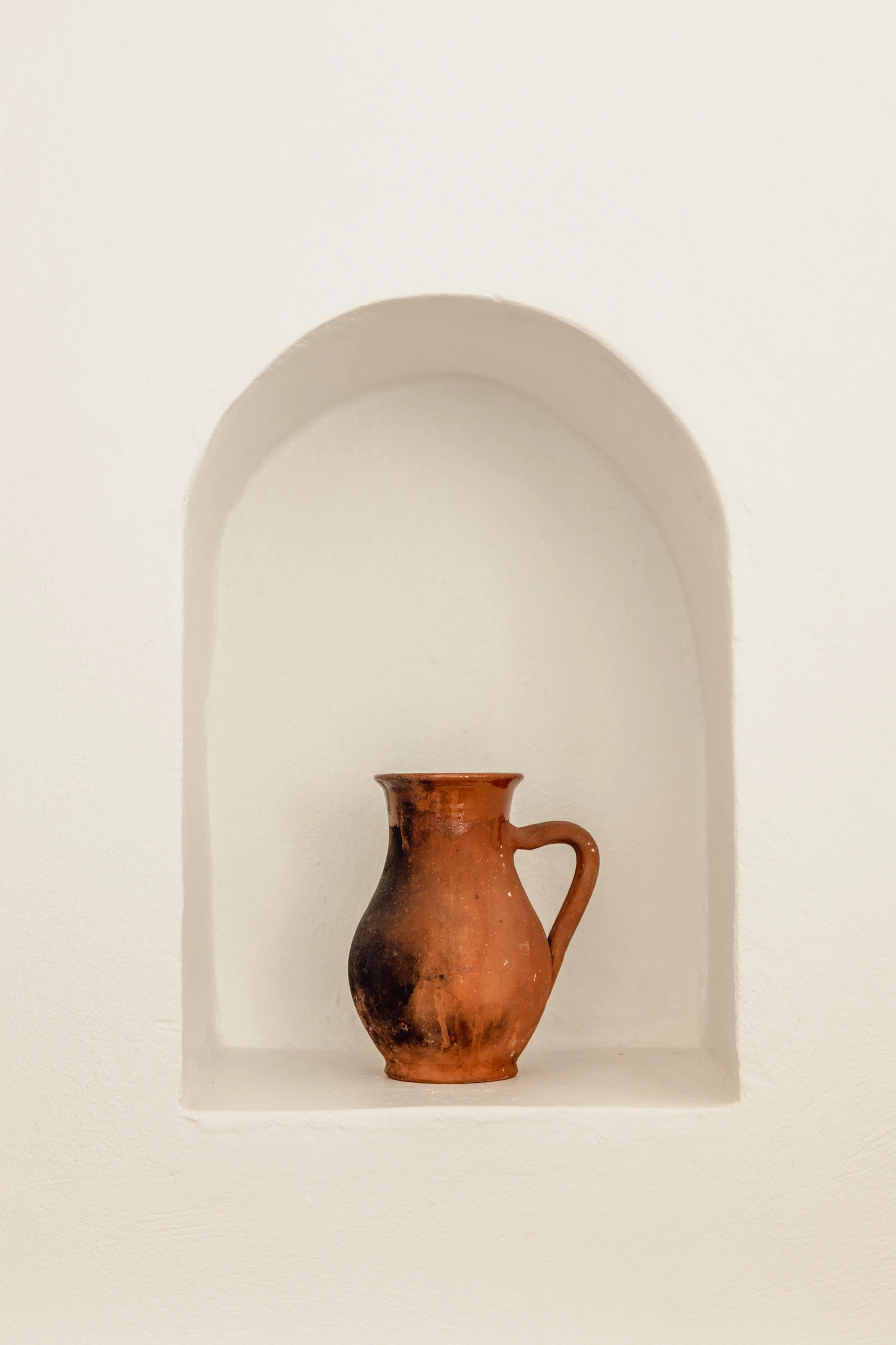 A small, ancient clay mug with a handle, displayed in a recessed wall niche painted white.