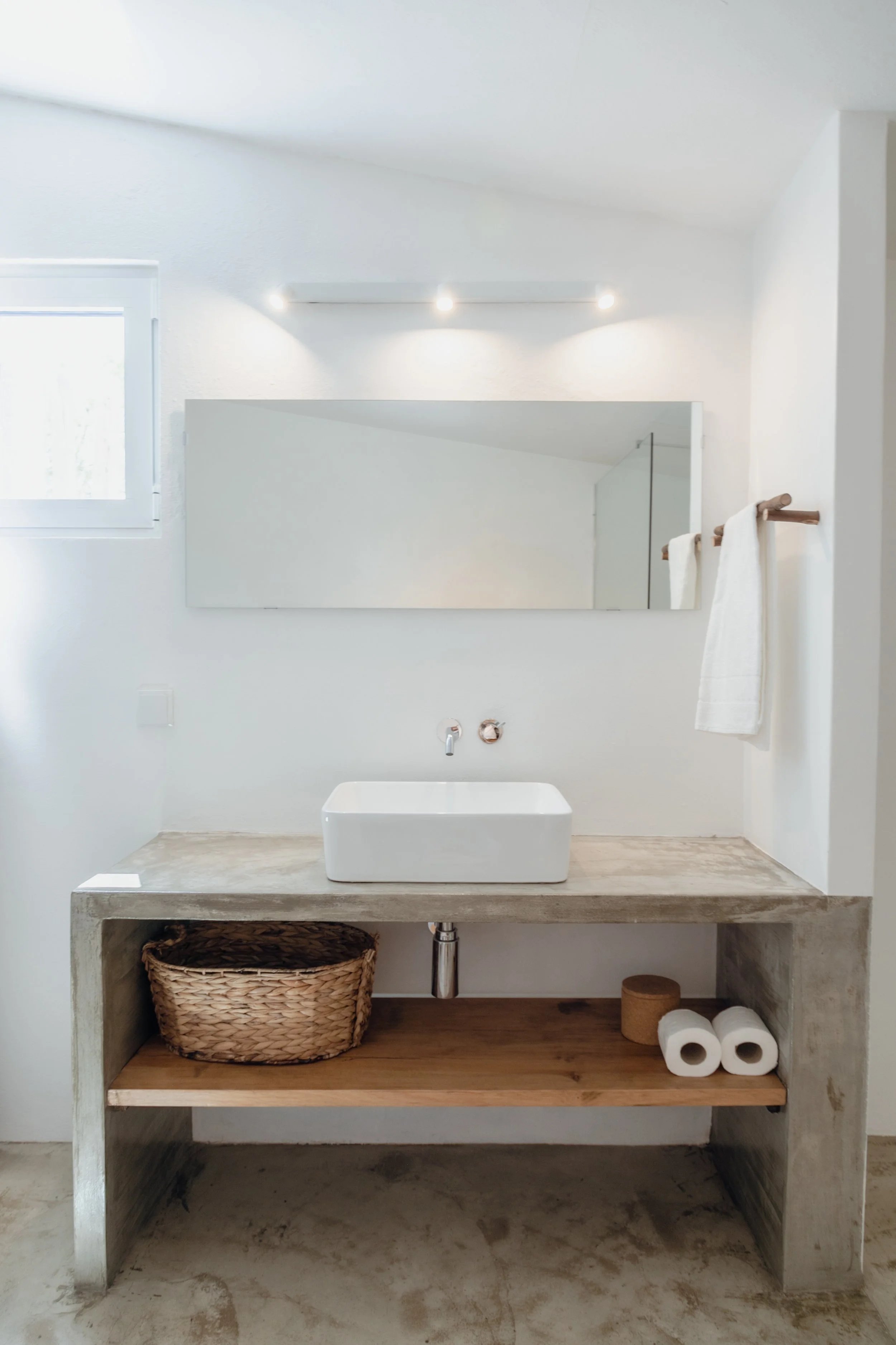 Minimalist bathroom with a rectangular vessel sink on a concrete countertop, a large wall mirror, towels hanging on a wooden rod, a small window, and a shelf with a woven basket and toilet paper rolls underneath.