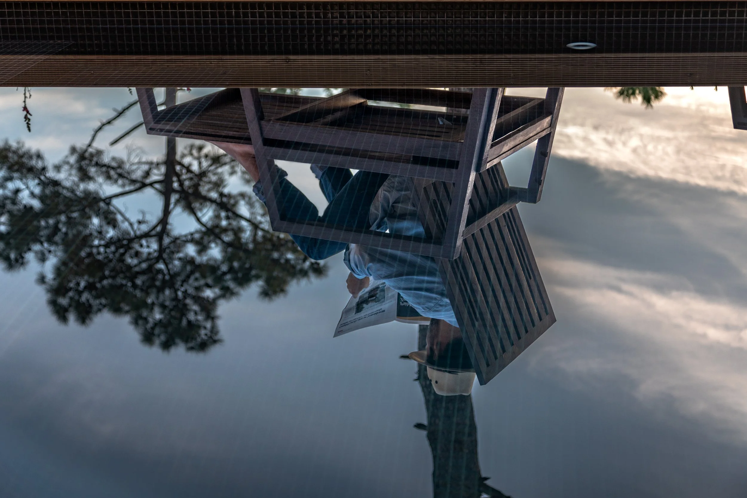 Reflection of a person sitting at an outdoor wooden table, reading a newspaper, with a tree and cloudy sky reflected in a water surface.