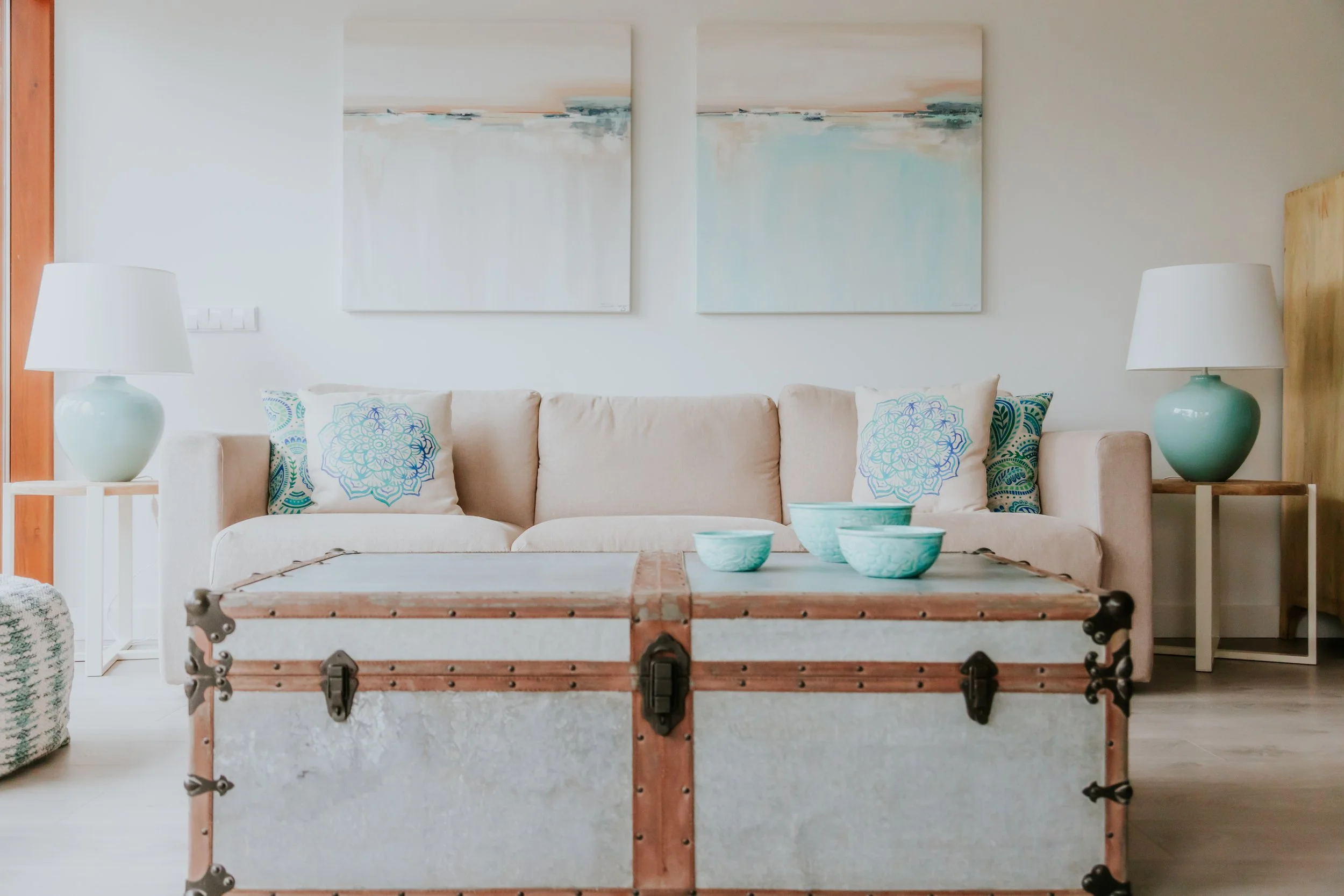 Living room with a beige sofa, two side tables with teal lamps, abstract wall art, decorative pillows, and a vintage trunk in front