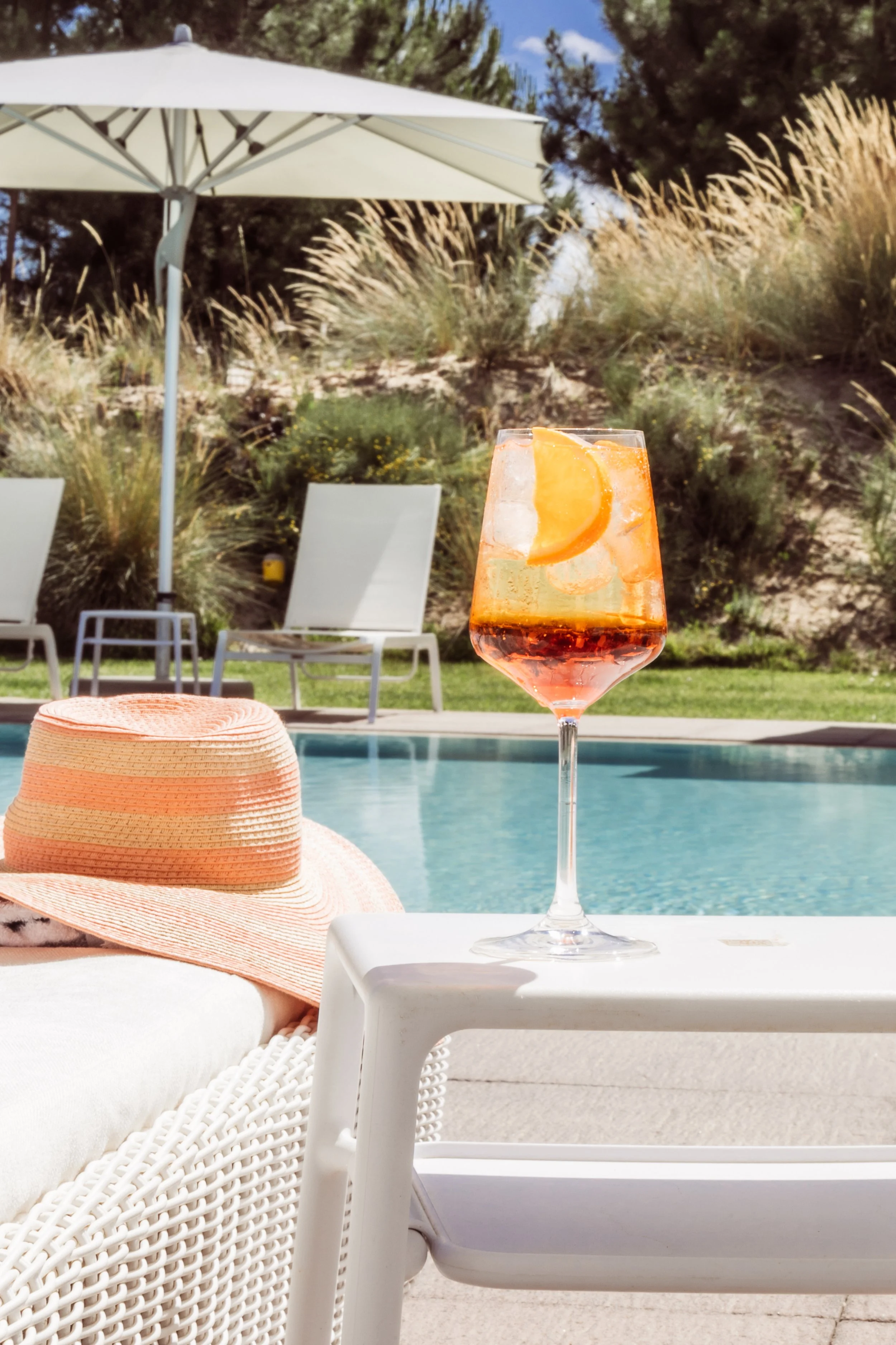 A poolside scene with a glass of orange-infused wine or cocktail, a straw sun hat on a white lounge chair, and a large white umbrella with lounge chairs and greenery in the background.
