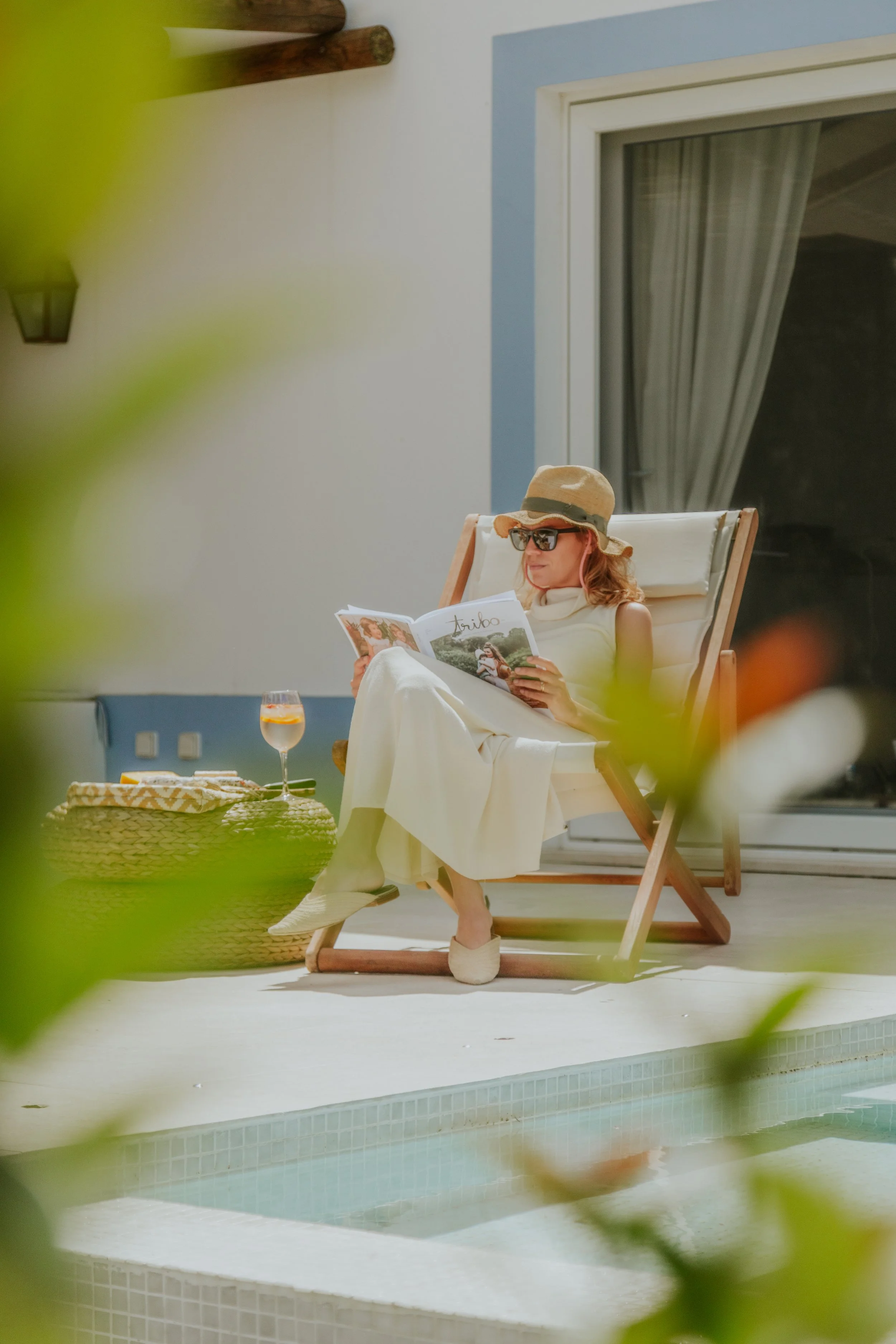 A woman relaxing in a lounge chair by a pool, wearing a wide-brimmed hat, sunglasses, and a white dress, reading a magazine, with a glass of rosé wine on a table nearby.