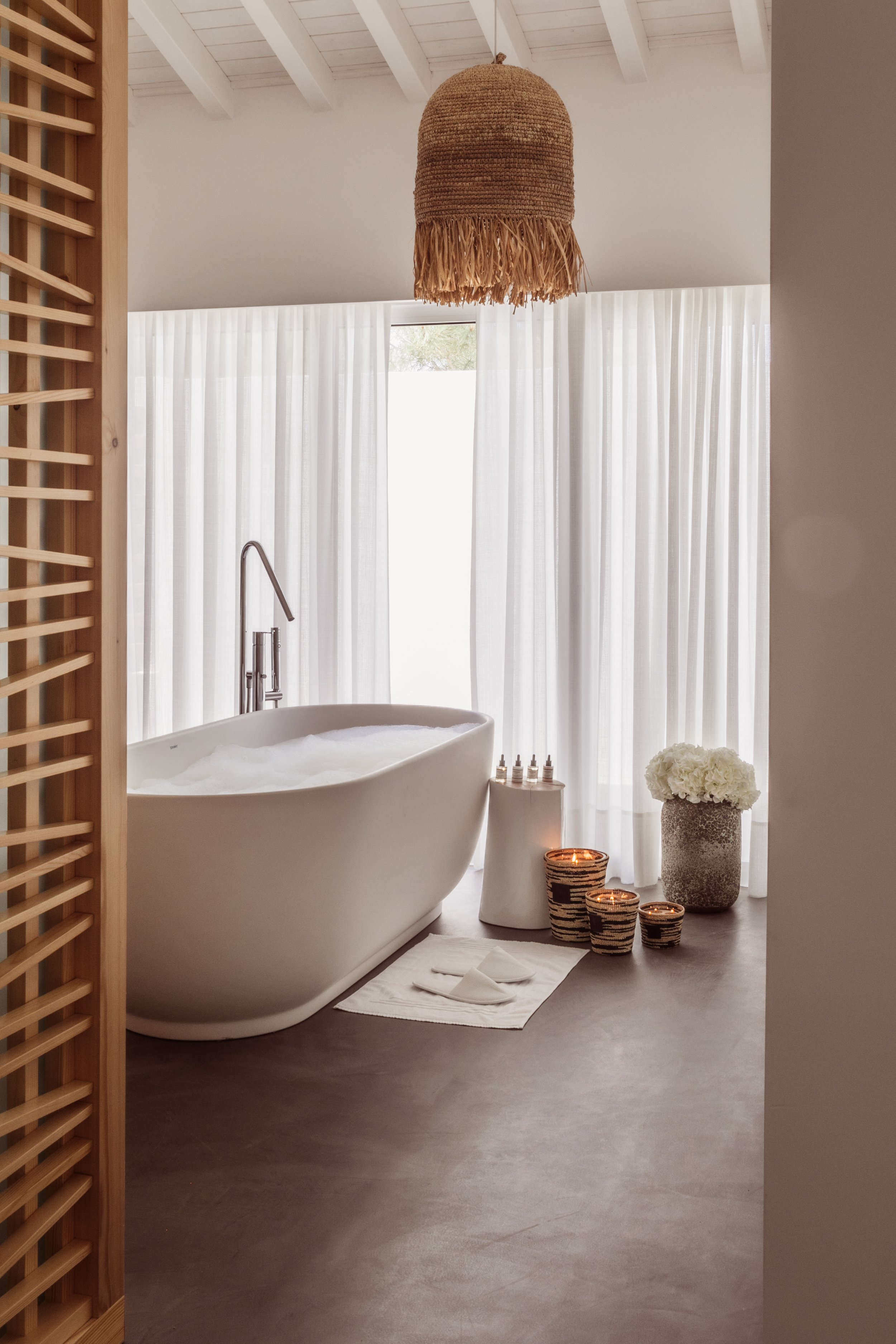 A modern bathroom with a freestanding bathtub filled with bubbles, surrounded by natural decor, white curtains, a woven hanging light fixture, and flowers in a large vase.