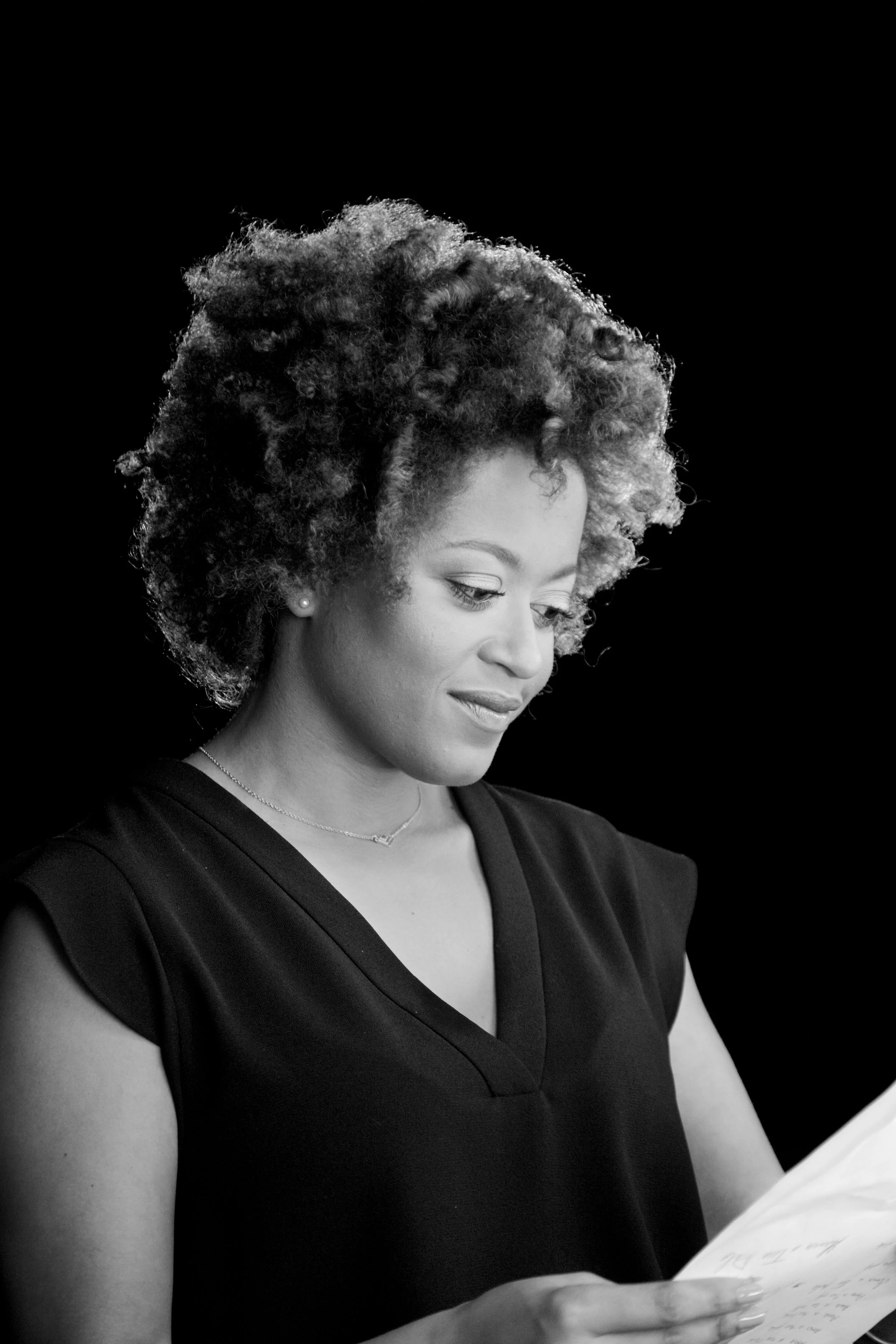 Black and white portrait of a woman reading a document, smiling gently, with curly hair, wearing a sleeveless top and a necklace.