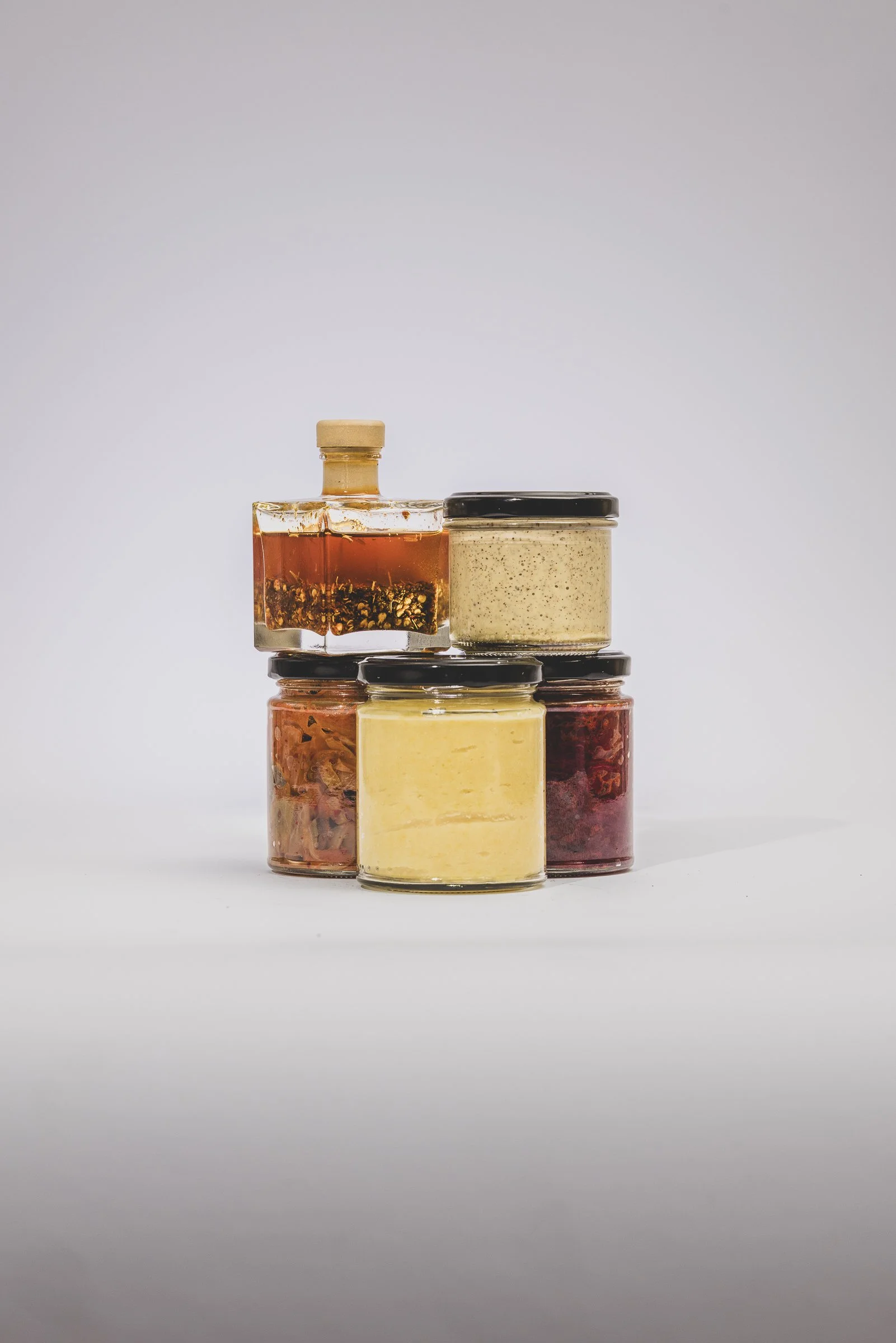A stack of five glass jars with various spices and condiments against a plain background.