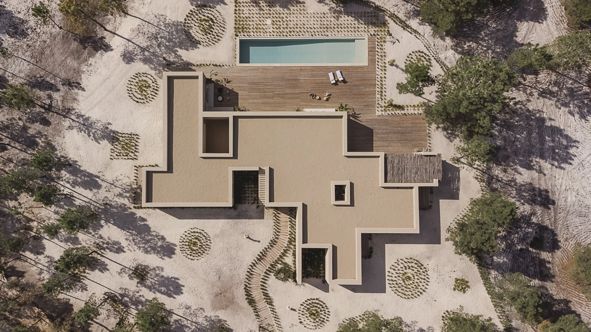 Aerial view of a modern house with a swimming pool on a sandy landscape, surrounded by trees and landscaping.