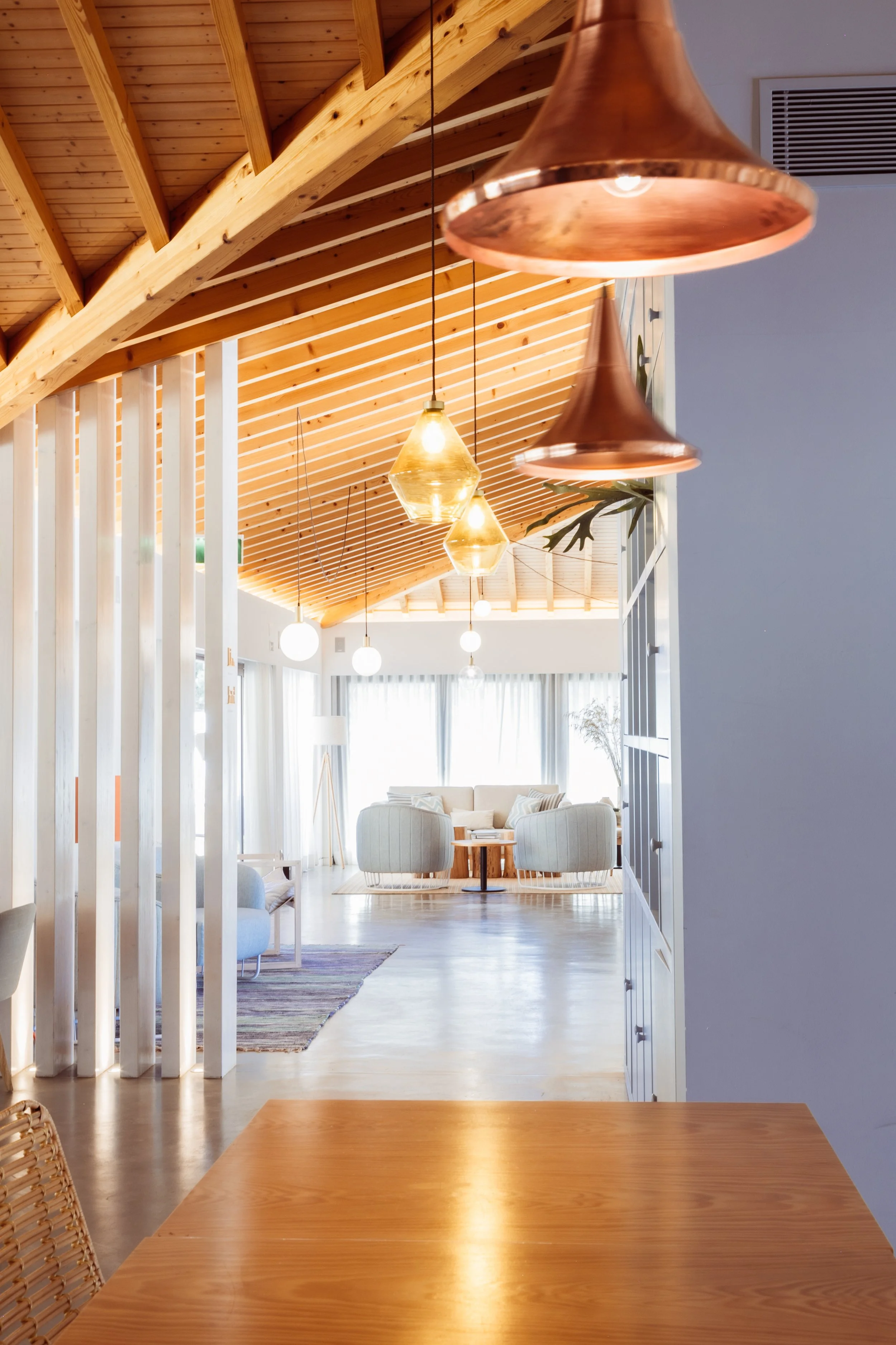 Modern interior living room with wooden vaulted ceiling, hanging pendant lights, and natural light coming through large windows with curtains.