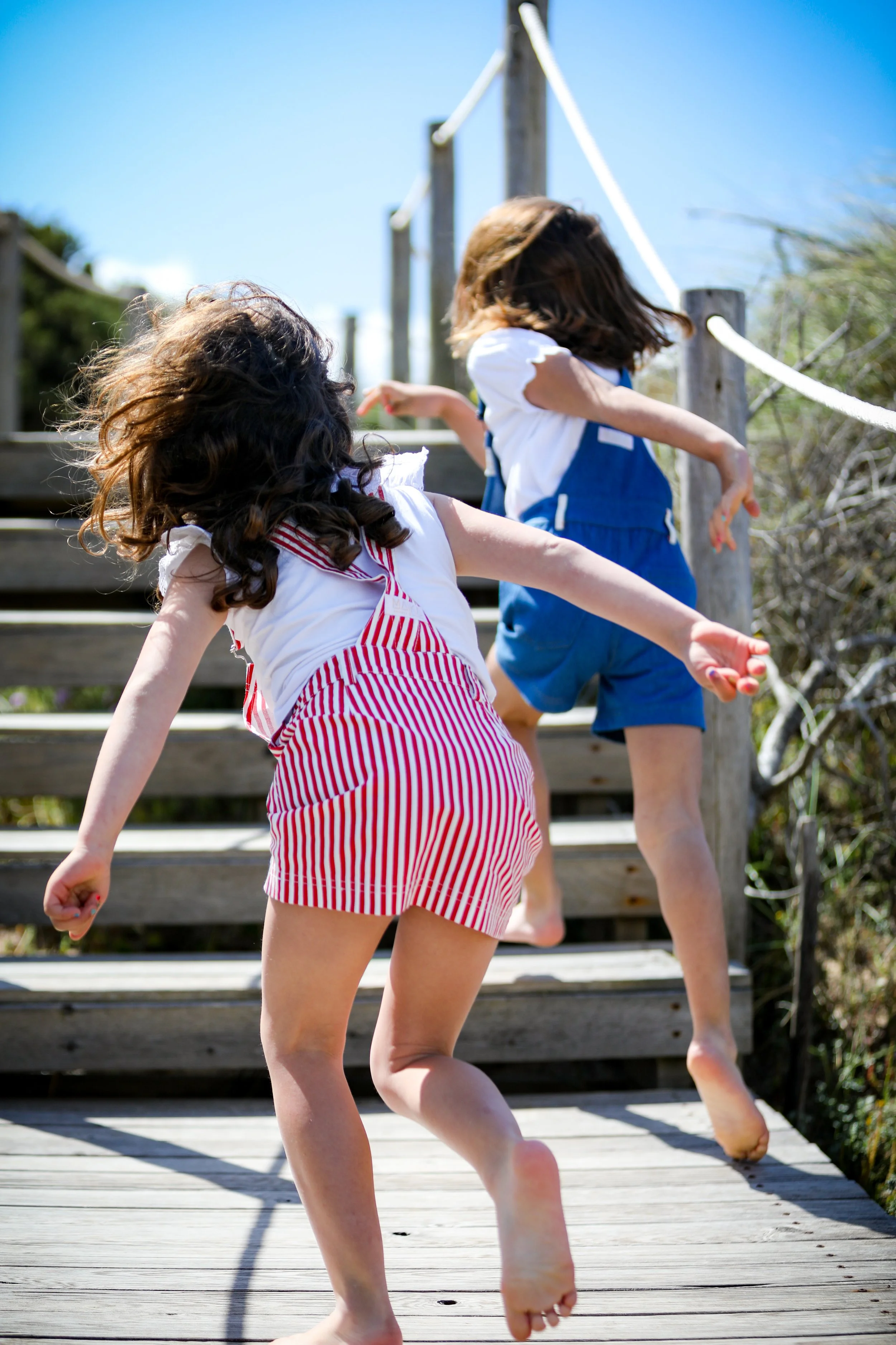 Two children playing on wooden stairs outdoors on a sunny day, one girl in red and white striped shorts and the other in blue overalls, emphasizing carefree childhood fun.