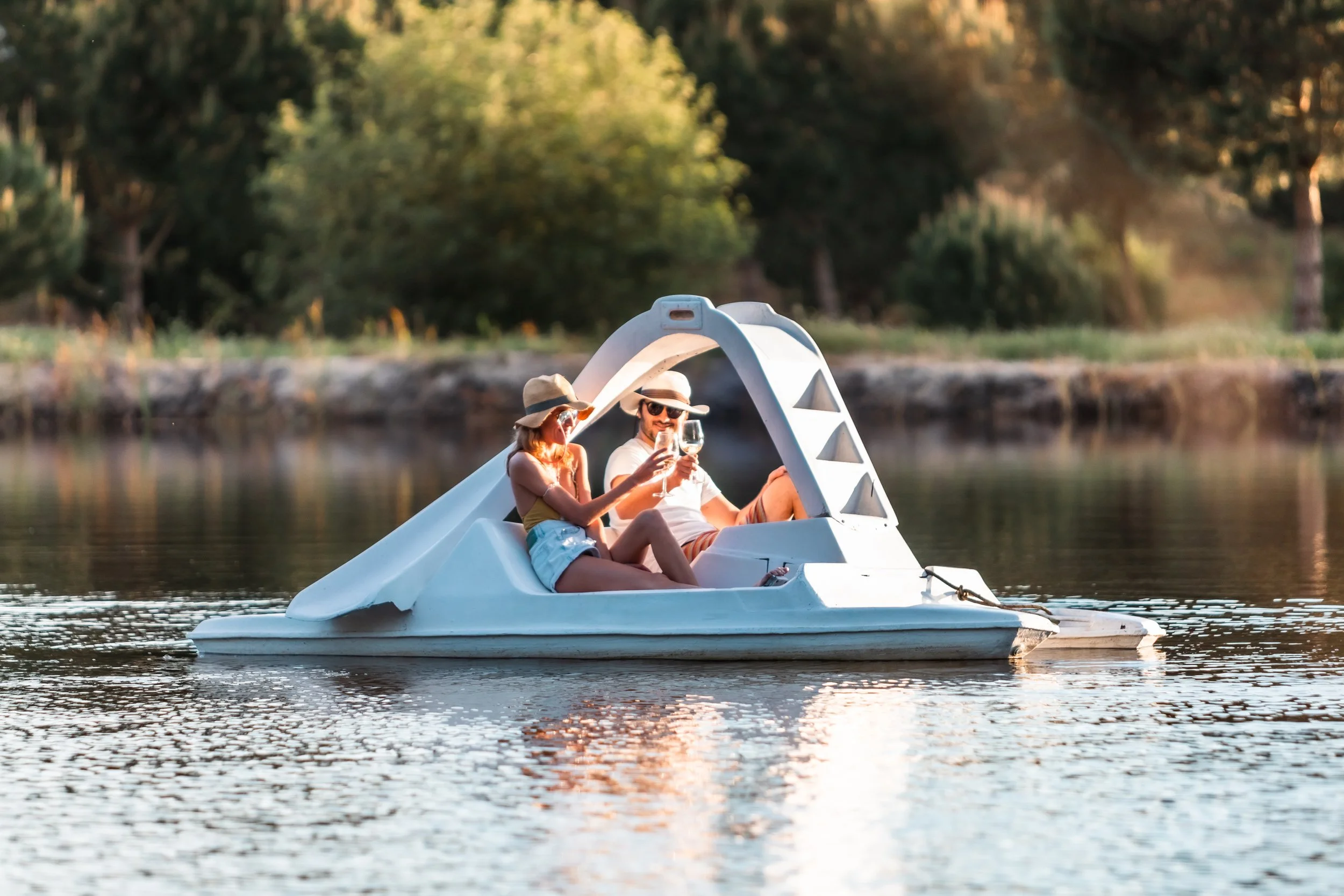 A couple in swimsuits and hats enjoying a boat ride on a lake during sunset, raising glasses of wine toward each other.