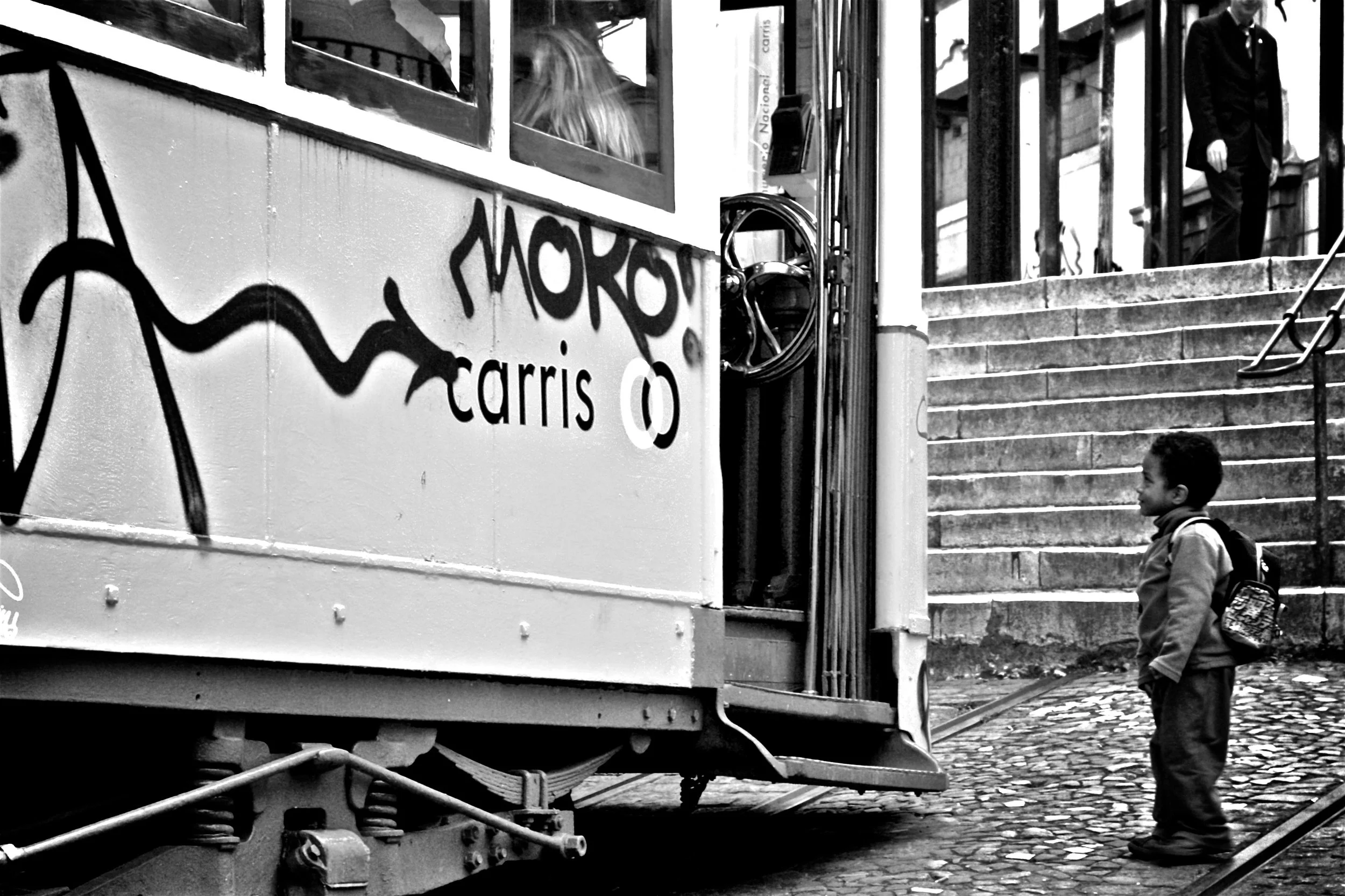 A young boy with a backpack waiting near a tram with graffiti on its side, on cobblestone pavement next to a staircase.