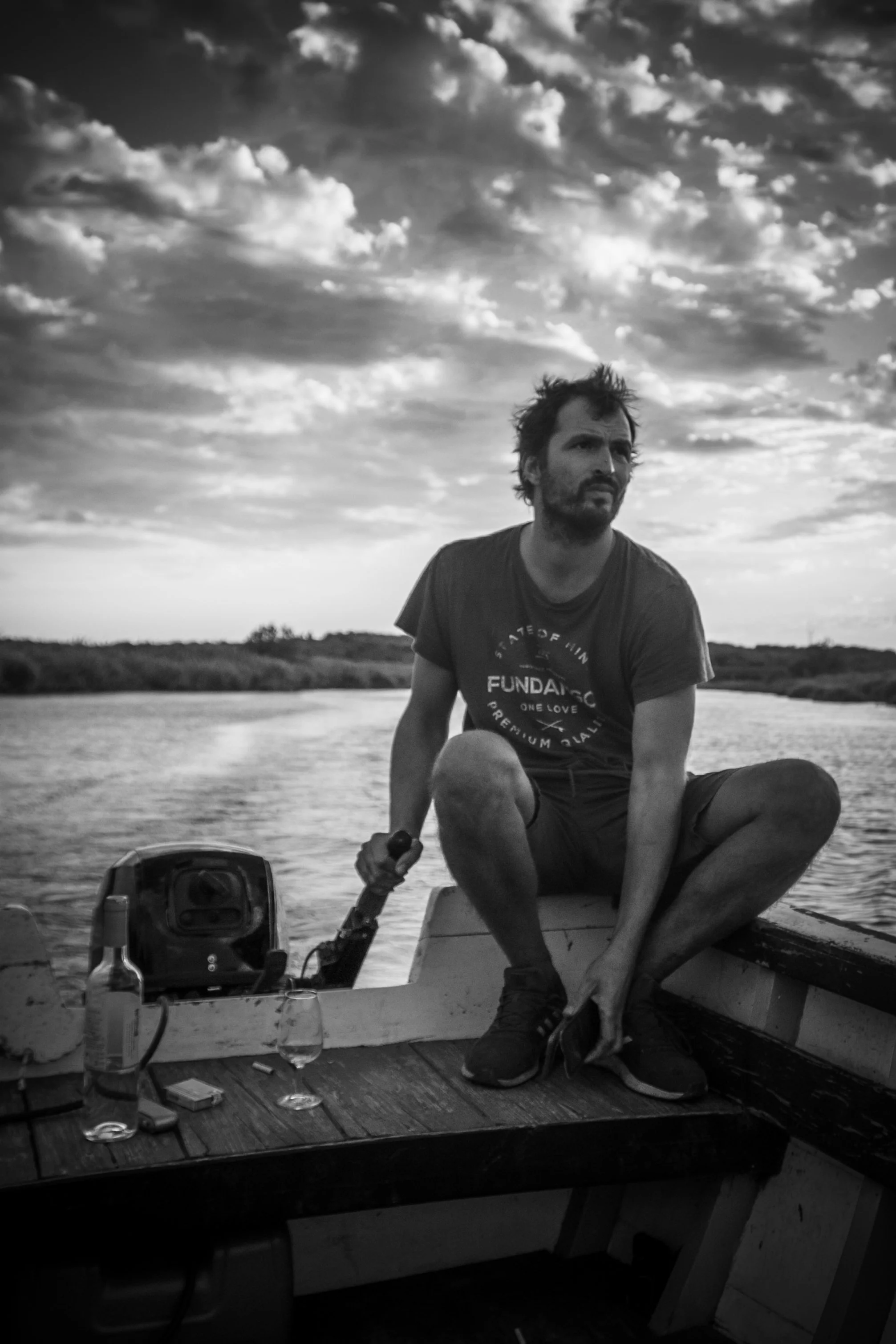 A man with disheveled hair and beard sitting on the edge of a small boat, holding a motorboat handle, with a river and cloudy sky in the background. There are bottles, glasses, and a phone on the boat's surface.