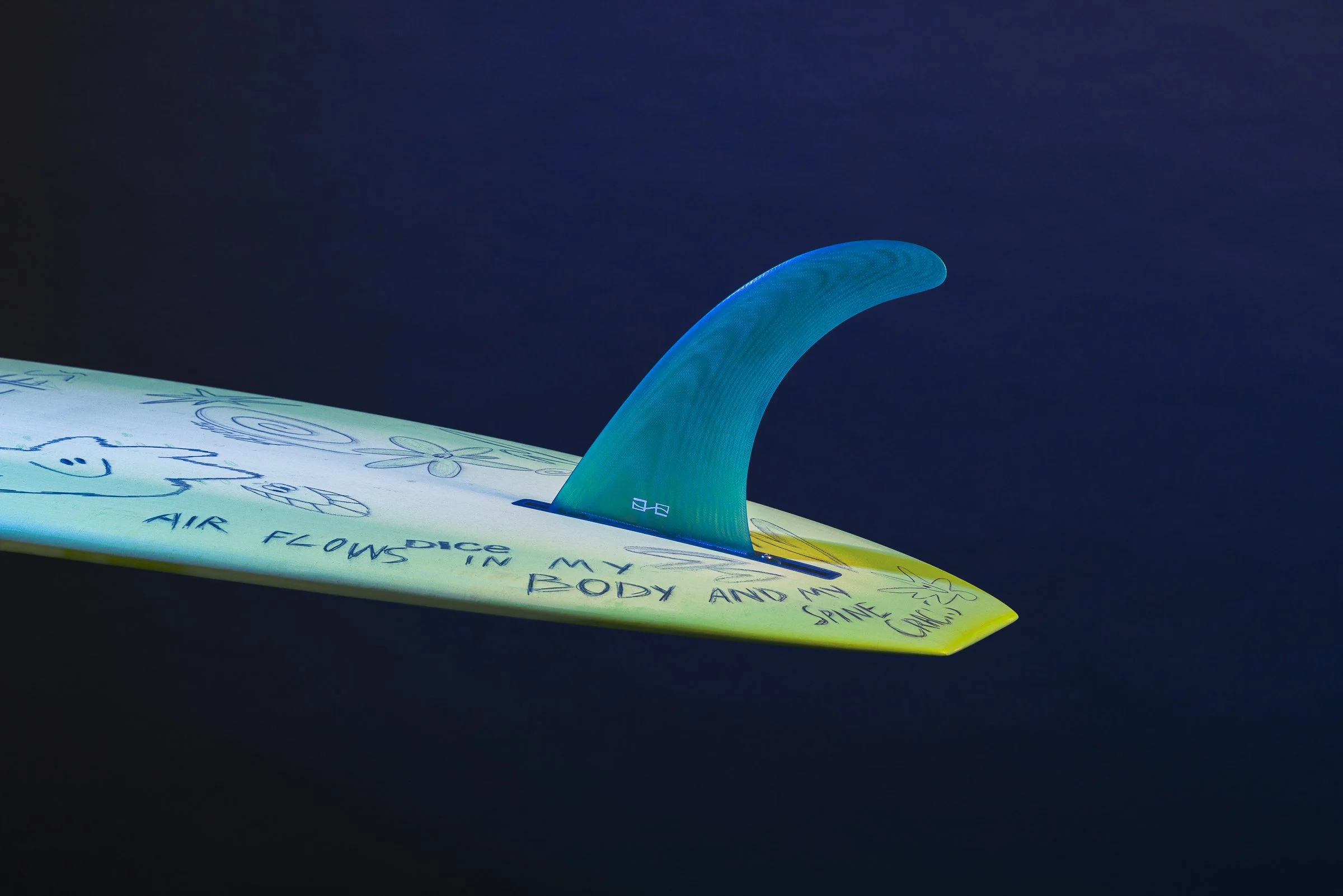 A surfboard with a dorsal fin attached, decorated with drawings and writing, floating against a dark background.