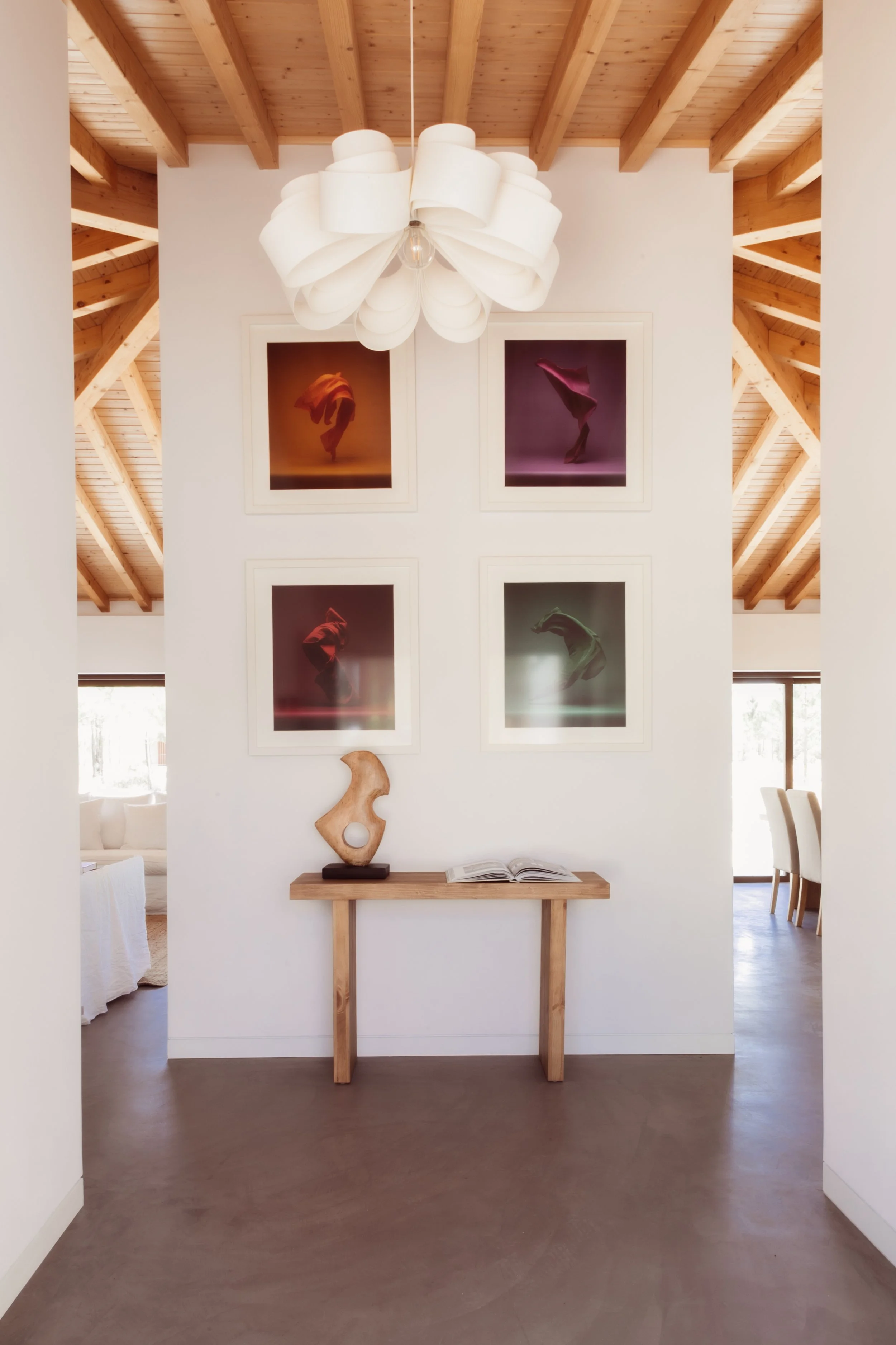 Interior view of a modern home with a ceiling lamp, four framed colorful artistic photographs on a white wall, and a small wooden console table with a decorative sculpture and an open book.
