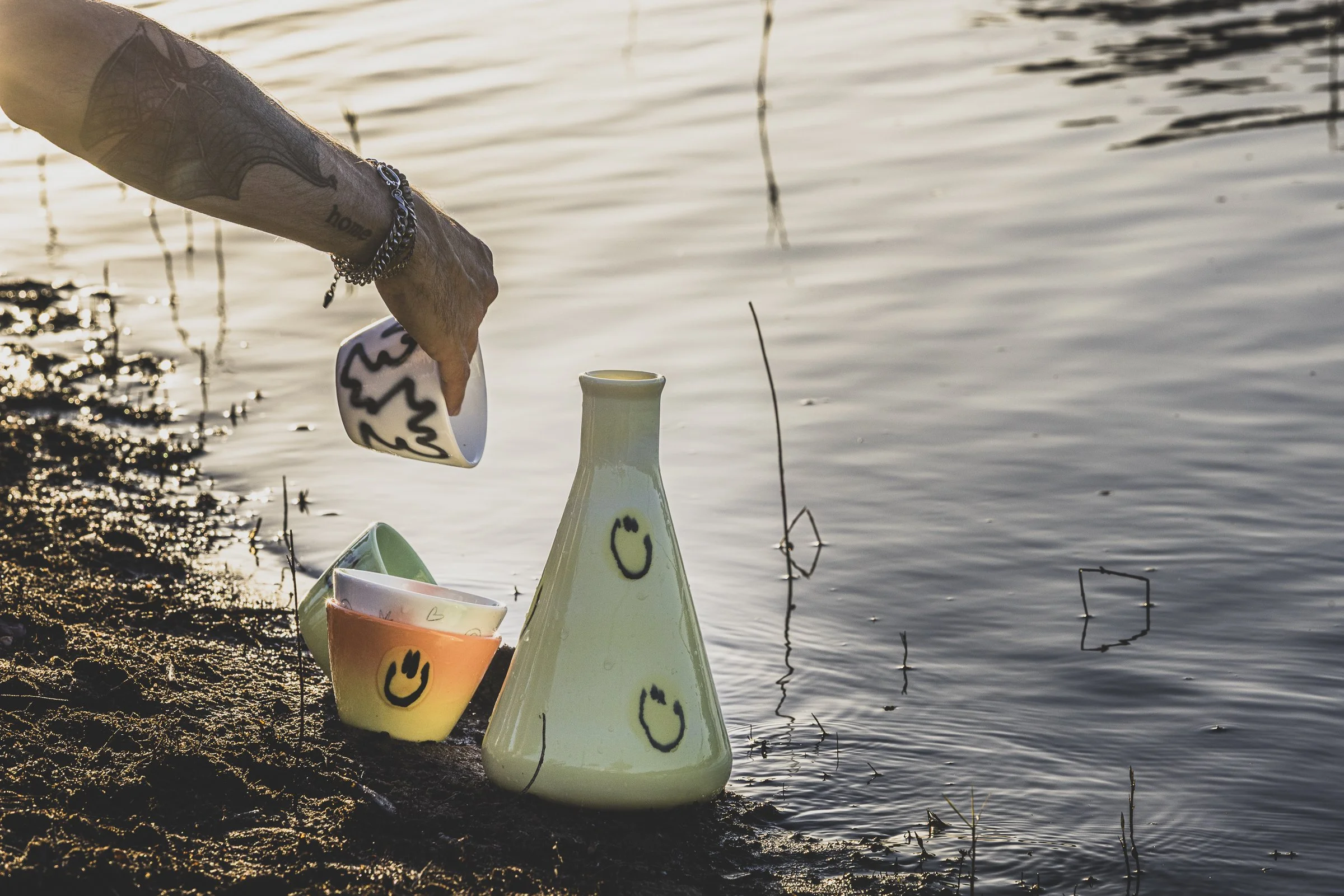 A person is placing a mug into a ceramic vessel shaped like a cone, with smiley faces drawn on both. There are two other colorful mugs nearby, all on the muddy shoreline by a calm body of water at sunset.