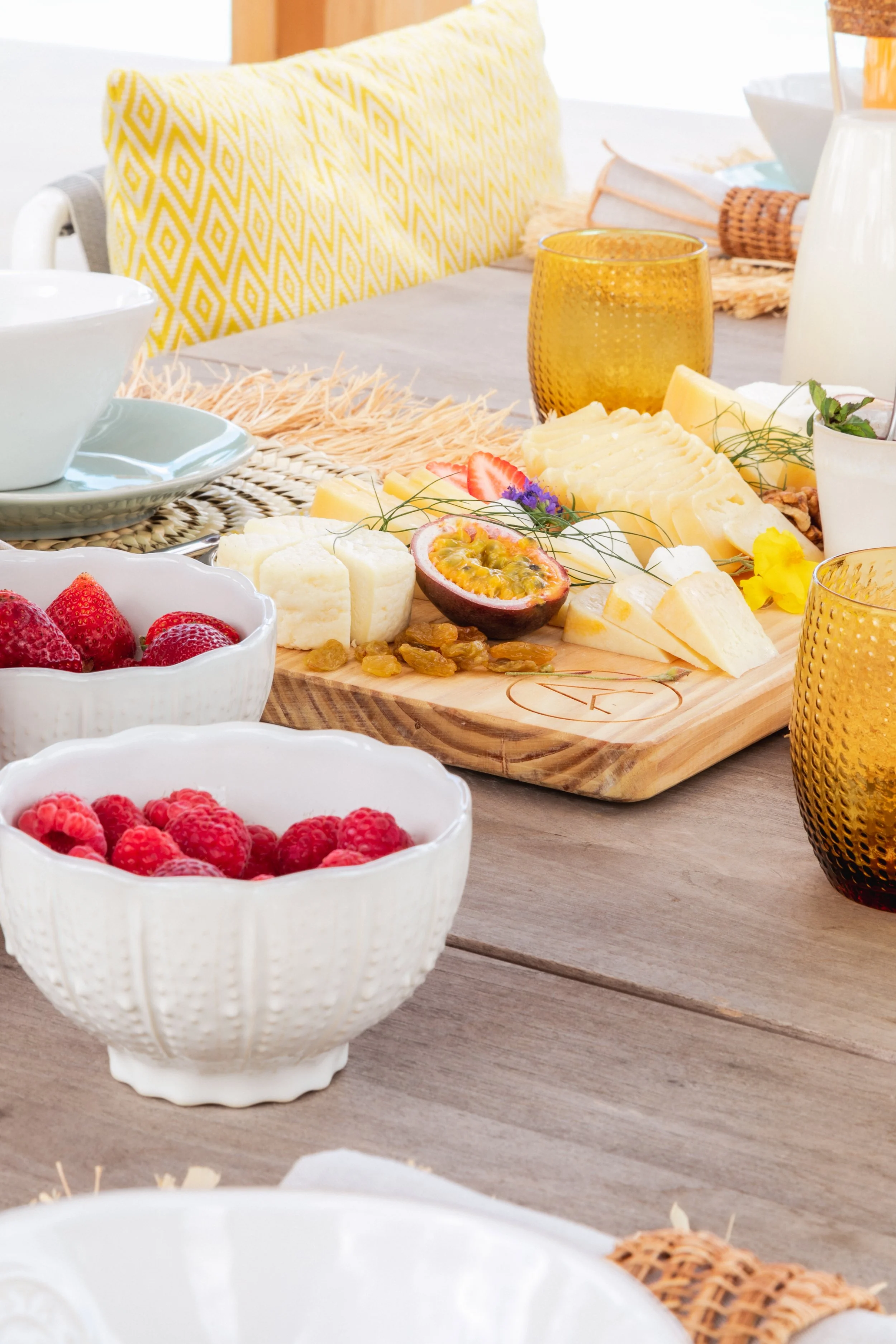 A breakfast spread including bowls of strawberries and raspberries, a cheese platter with assorted cheeses, sliced fruits, and nuts, on a dining table with amber glasses, plates, and a yellow pillow in the background.
