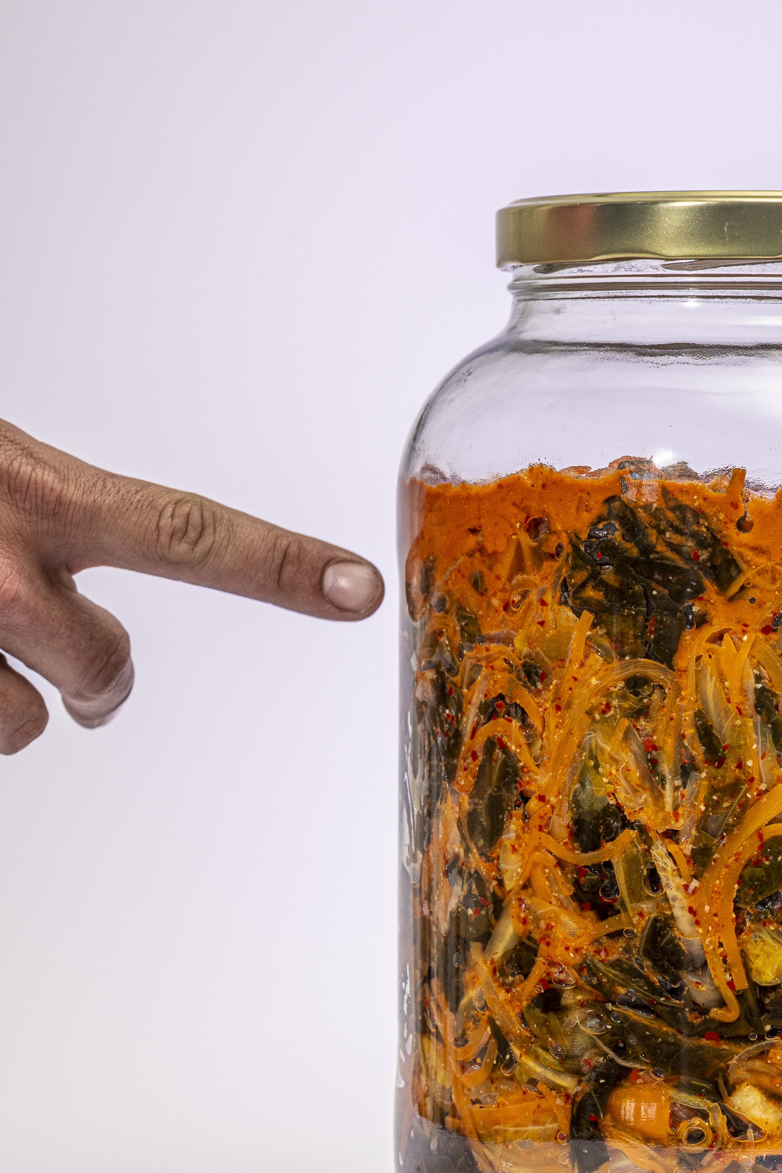 A finger pointing at a glass jar filled with spicy pickled vegetables.