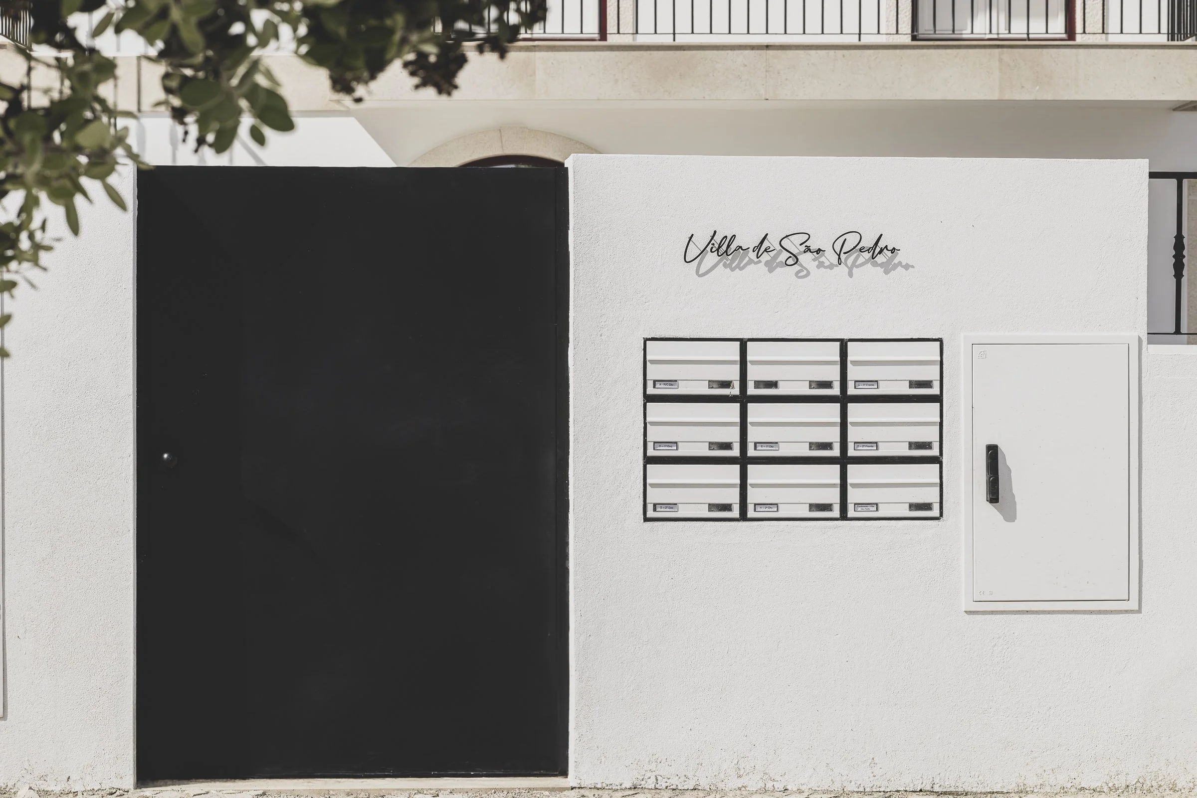 A modern apartment building entrance with a black gate on the left, white mailboxes in the center, and a white utility box on the right. The building is labeled 'Villa de São Pedro' written in black cursive above the mailboxes. There are trees or bus