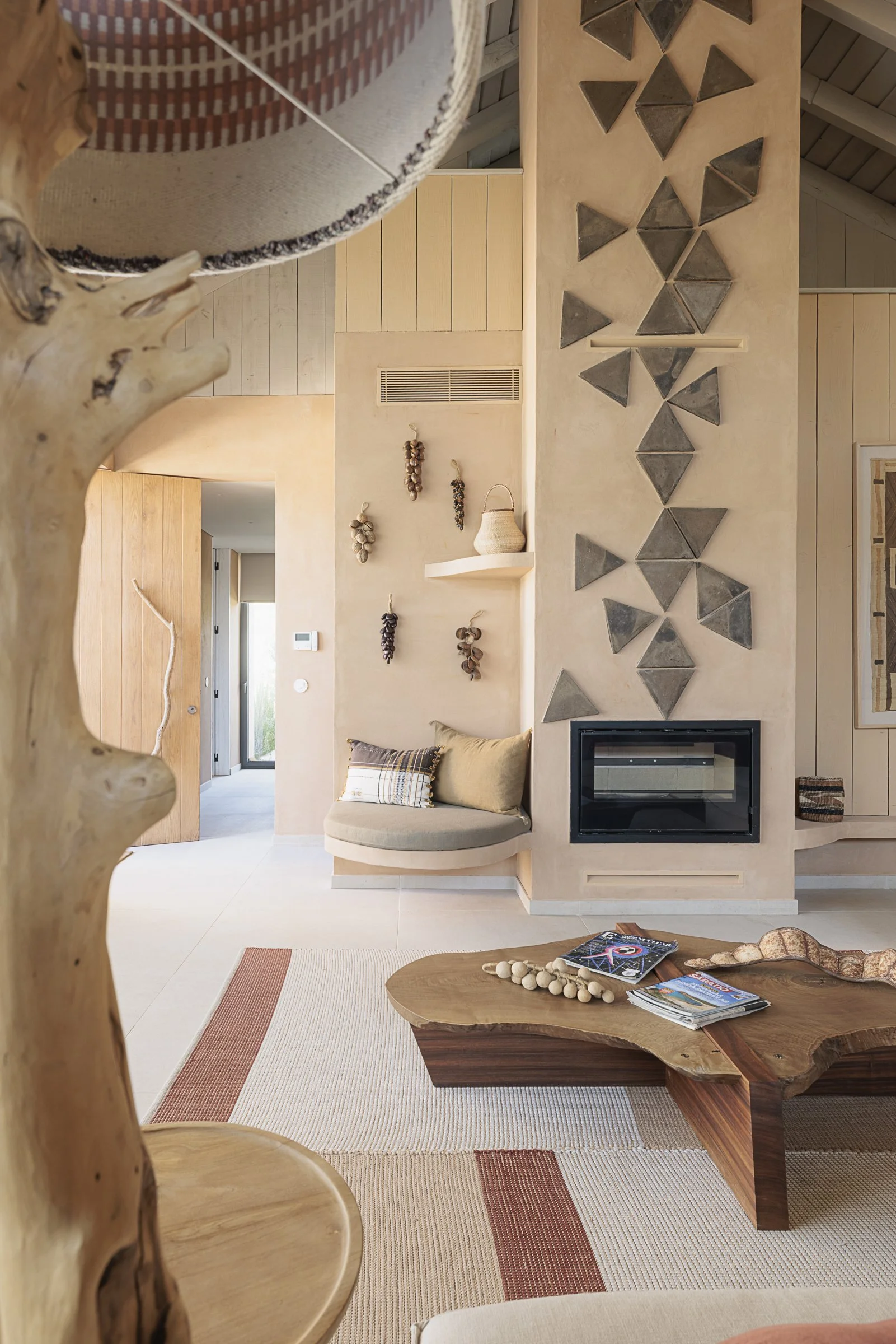 Interior of a modern living room with a natural wood coffee table, a cozy niche with a cushion and pillows, a fireplace, and decorative wall art including hanging garlic and a shell basket.