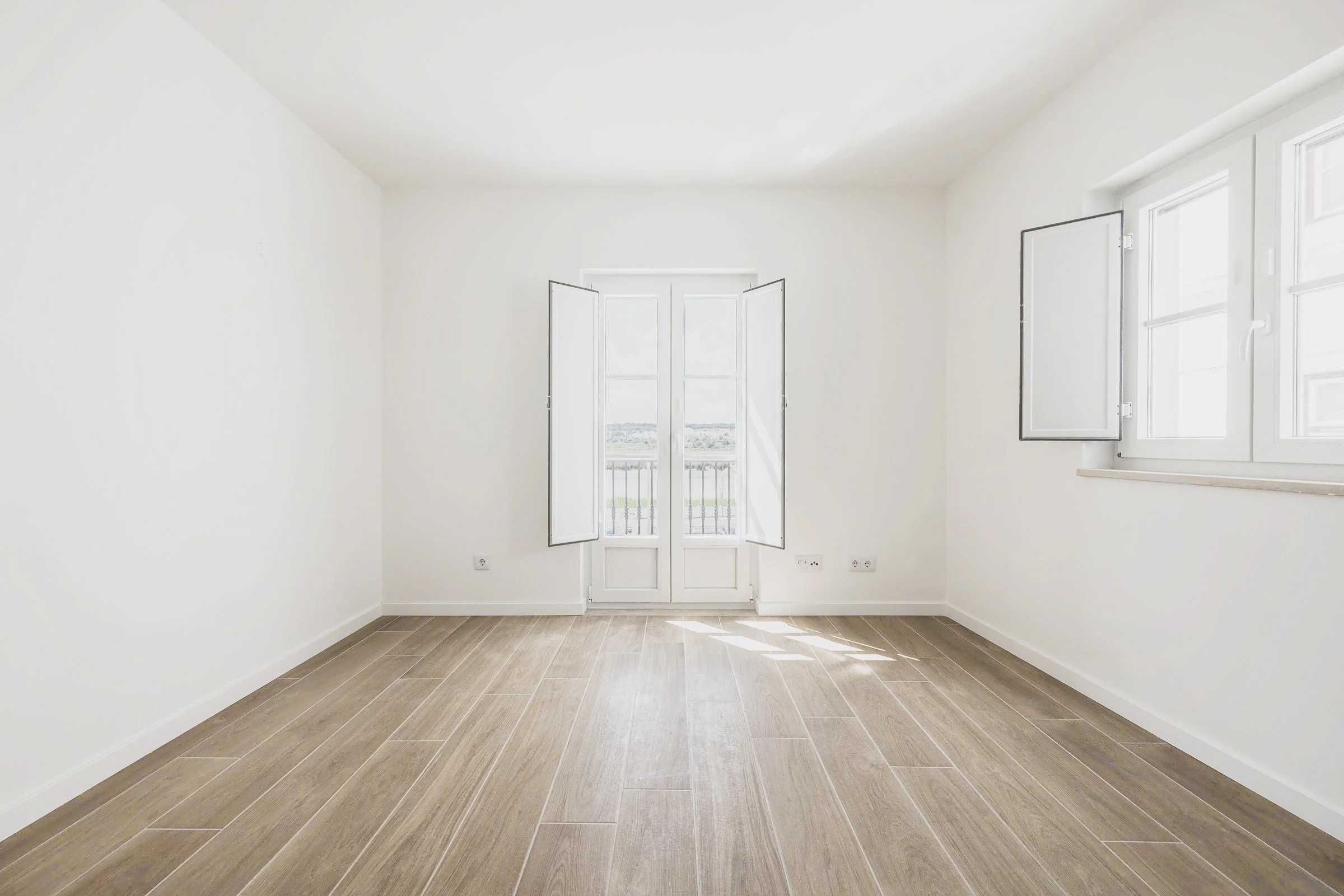 Empty room with white walls, wooden floor, and open windows with a small balcony outside, allowing sunlight to brighten the room.