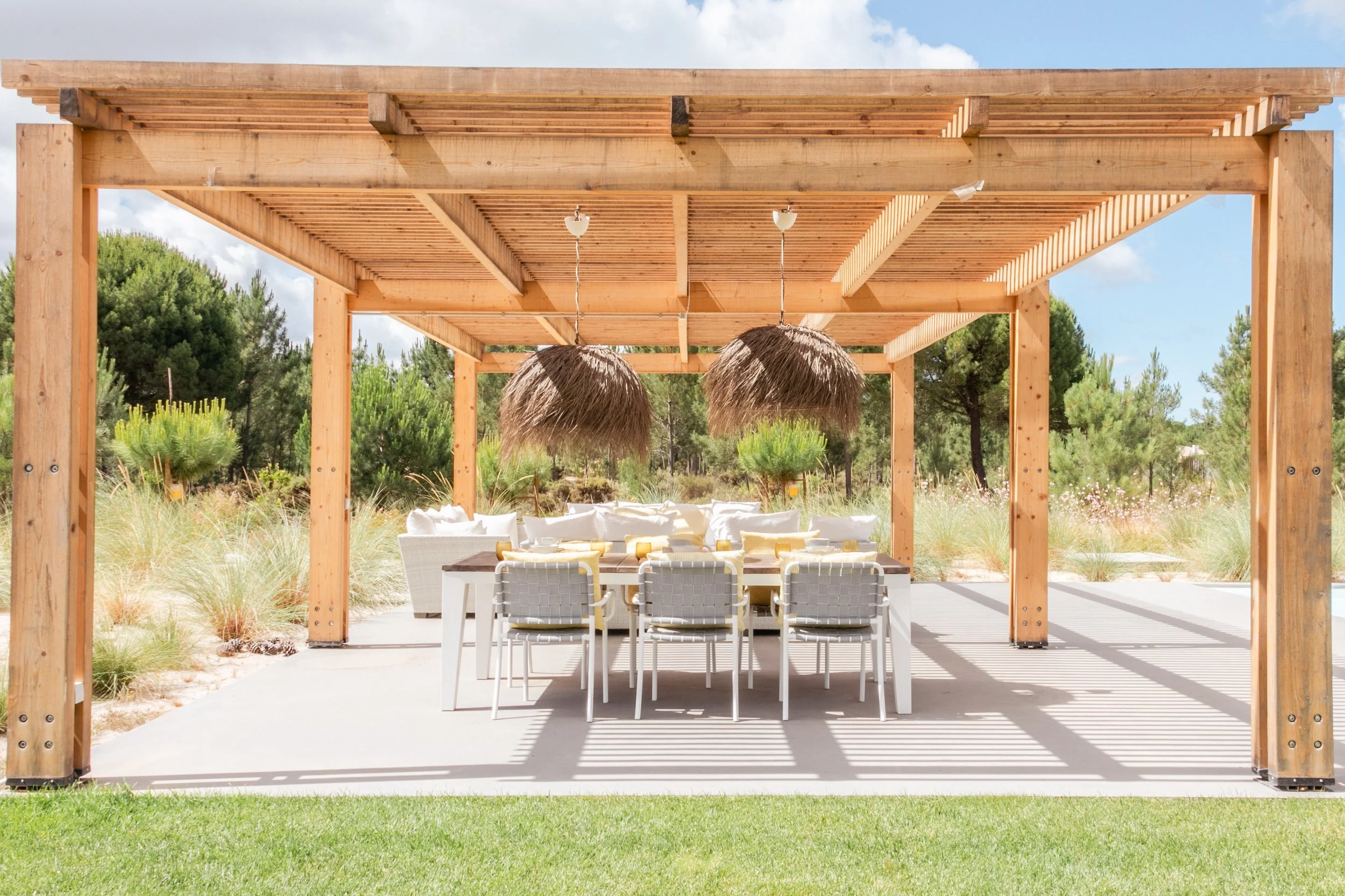 Outdoor patio with a wooden pergola, hanging straw pendant lights, a dining table with white chairs, surrounded by grassy landscape and trees.