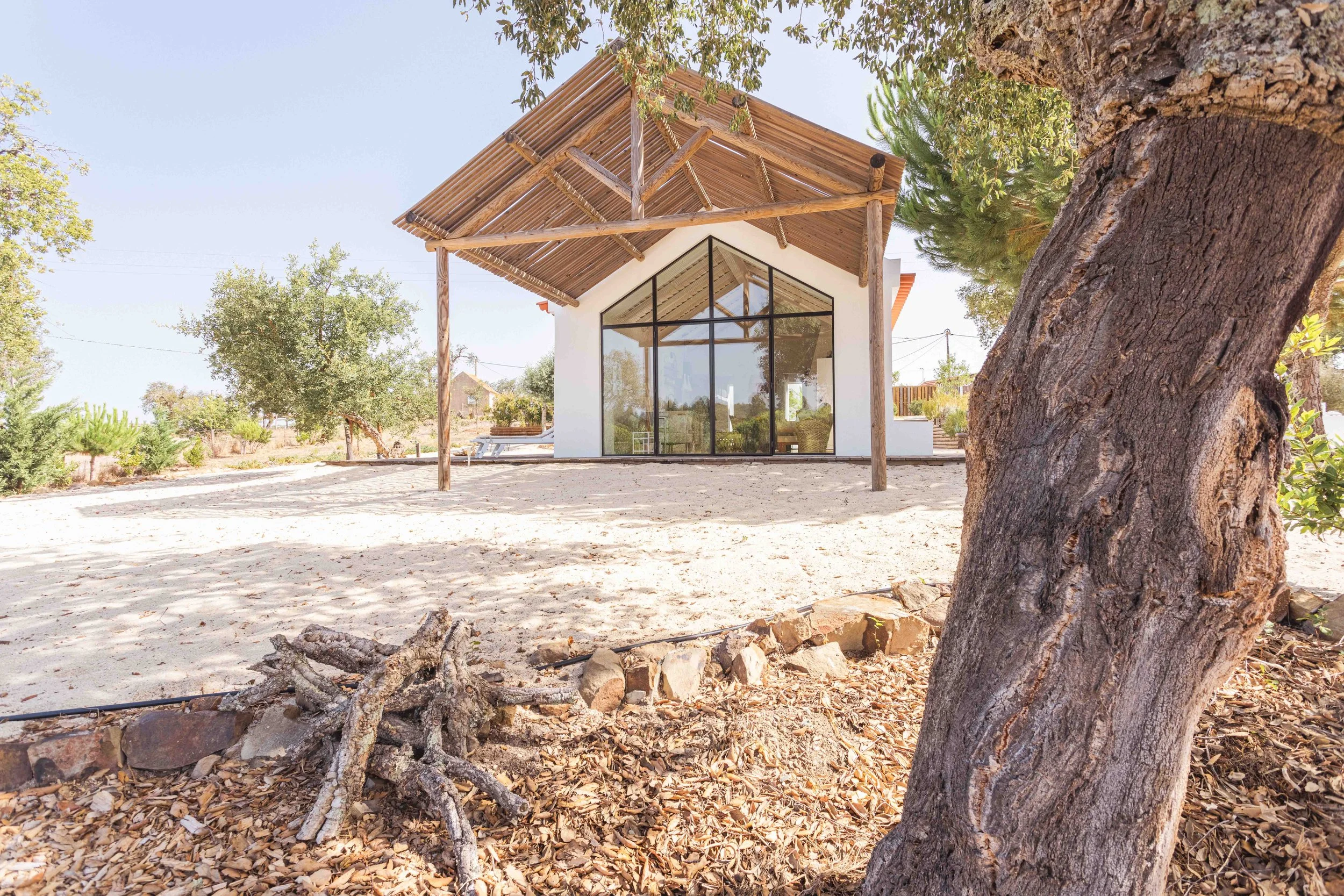 A small house with large glass windows and a wooden slat roof, situated on a sandy yard with trees and plants around.