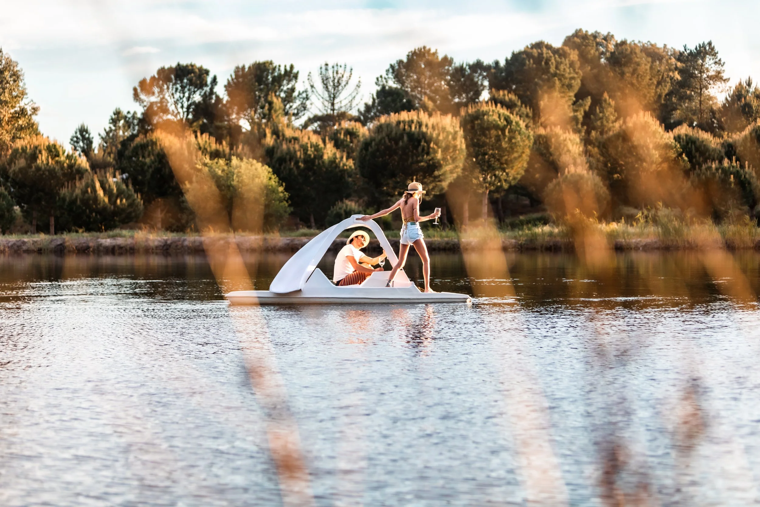 Two women enjoying a paddle boat ride on a lake during sunset, with trees in the background and reeds in the foreground.