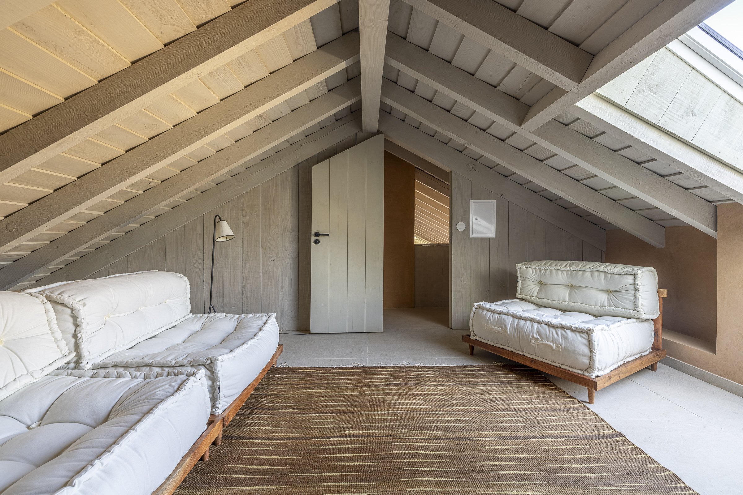 Attic living space with two white cushioned sofas and a striped brown rug, wooden walls, exposed wood ceiling beams, a small lamp, and a window on the sloped ceiling.