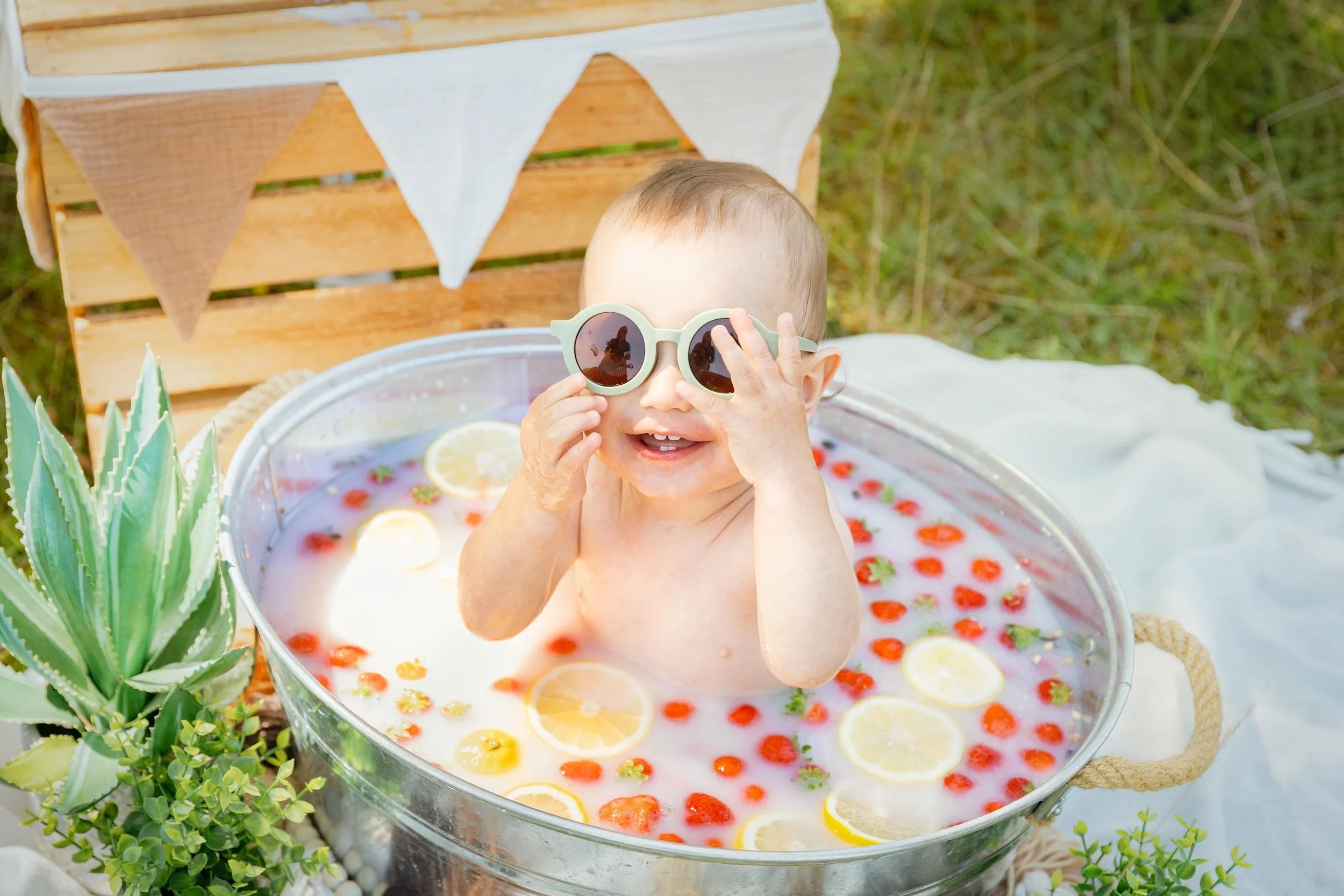 ein kleines Kind sitzt in einem Milchbad mit Zitronen und Erdbeeren darin. Das Baby hat eine Sonnenbrille auf und lacht in die Kamera. Das Foto wurde in einer Wiese aufgenommen