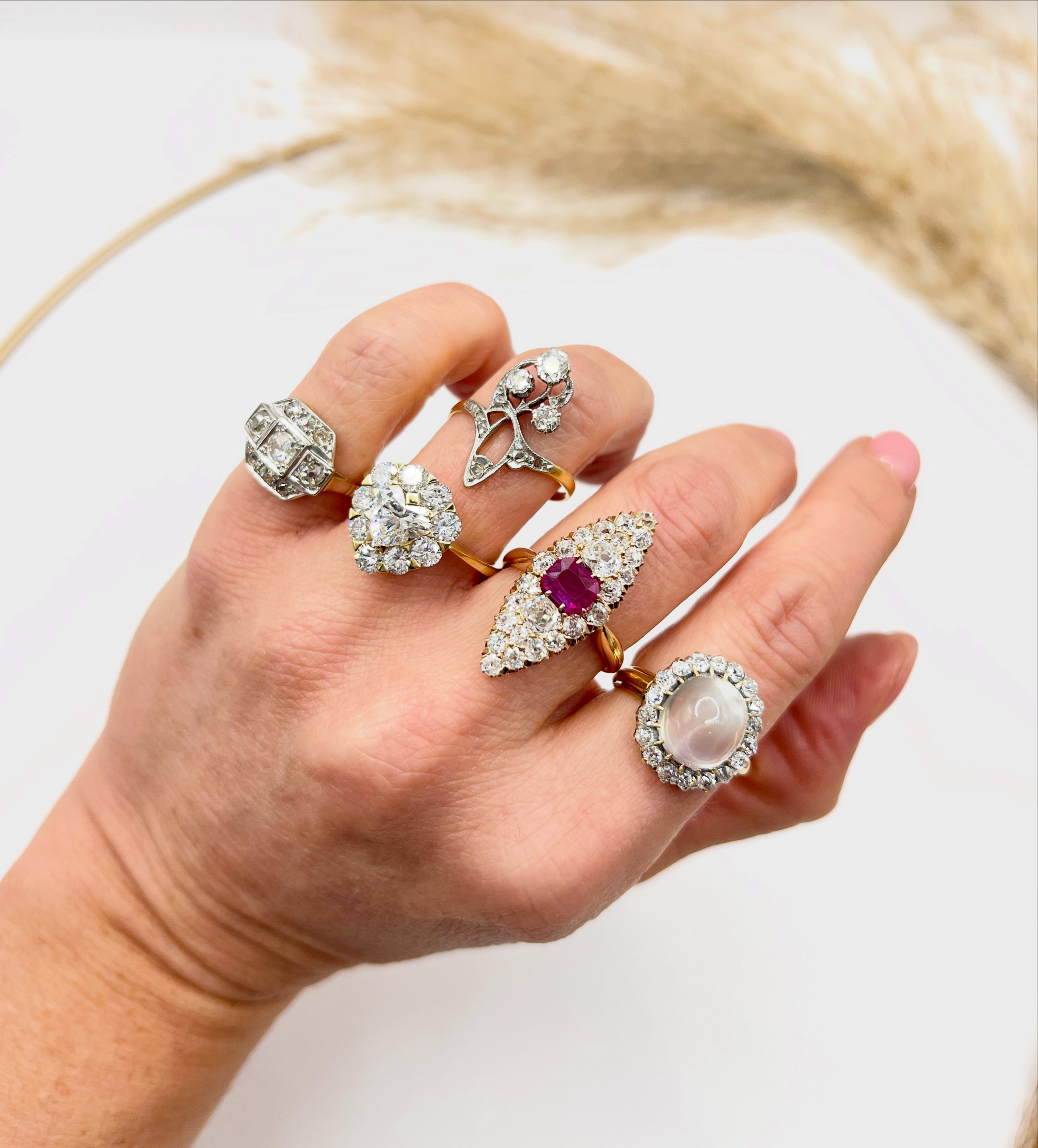 A hand with multiple rings, including a large pearl and diamond rings, jewelry with gemstones, and decorative designs, against a white background.