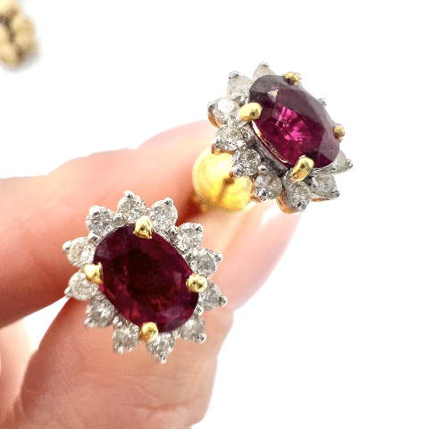 A pair of gold and diamond earrings with large oval ruby gemstones, held between a person's thumb and index finger.