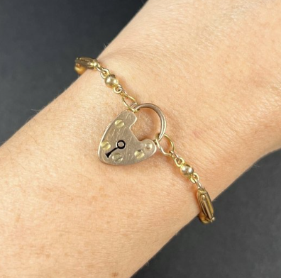 Gold bracelet with a heart-shaped padlock charm and a small rectangular charm on a person's wrist.