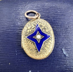 A gold oval-shaped pendant with intricate engraved patterns and a central blue star with a pearl in the middle, hanging from a metal loop, placed on a dark blue wooden surface.