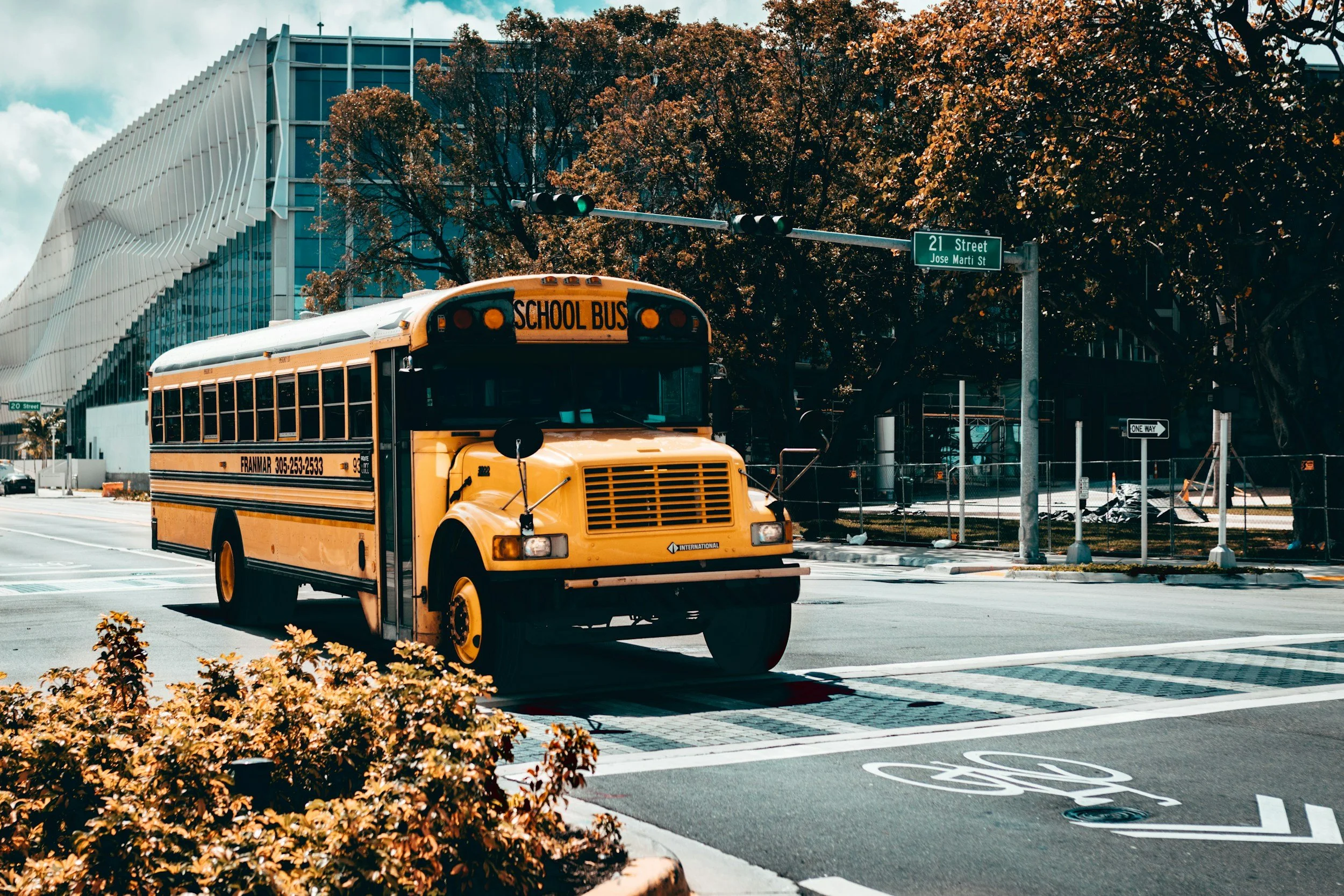 A yellow school bus driving on a city street, with trees and modern buildings in the background. There are street signs indicating 21st Street and Jose Marti Street, and a bicycle lane marked on the road.