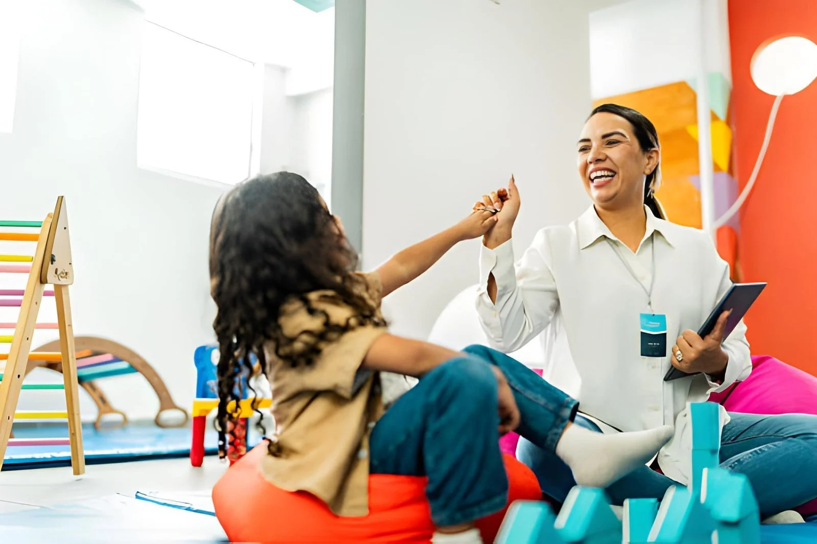 A woman smiling and holding a tablet interacts with a young girl in a colorful playroom.