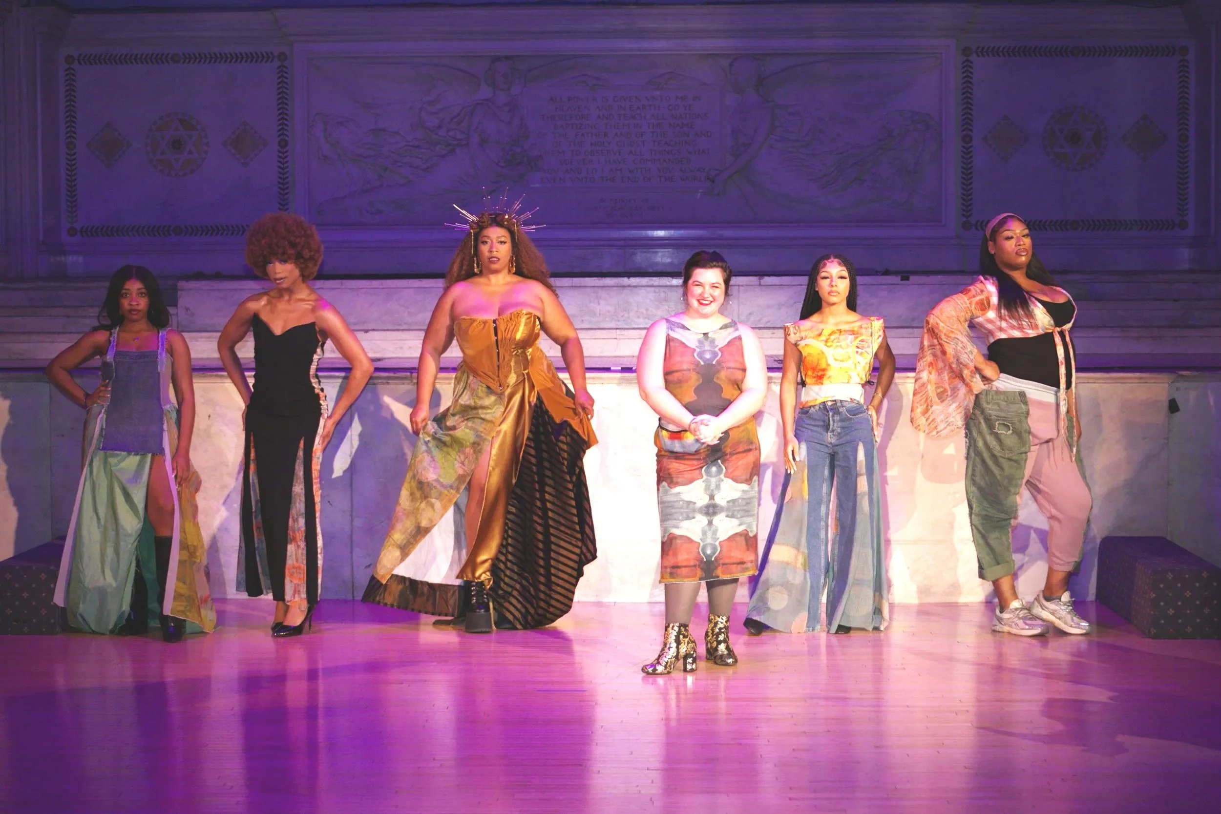 A diverse group of seven women standing on a stage, dressed in colorful and stylish outfits, posing confidently.