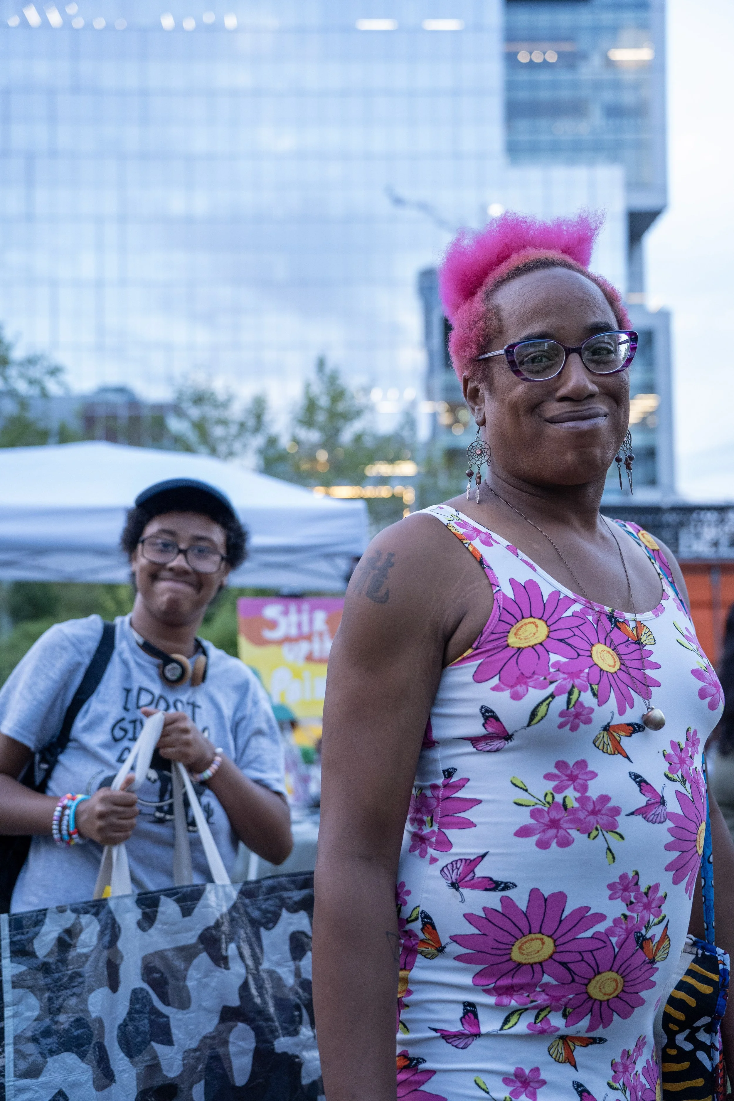 A woman with pink hair and glasses wearing a floral dress with butterflies stands in the foreground, smiling. A person with glasses, headphones around their neck, and a gray t-shirt stands in the background near a white tent, smiling.