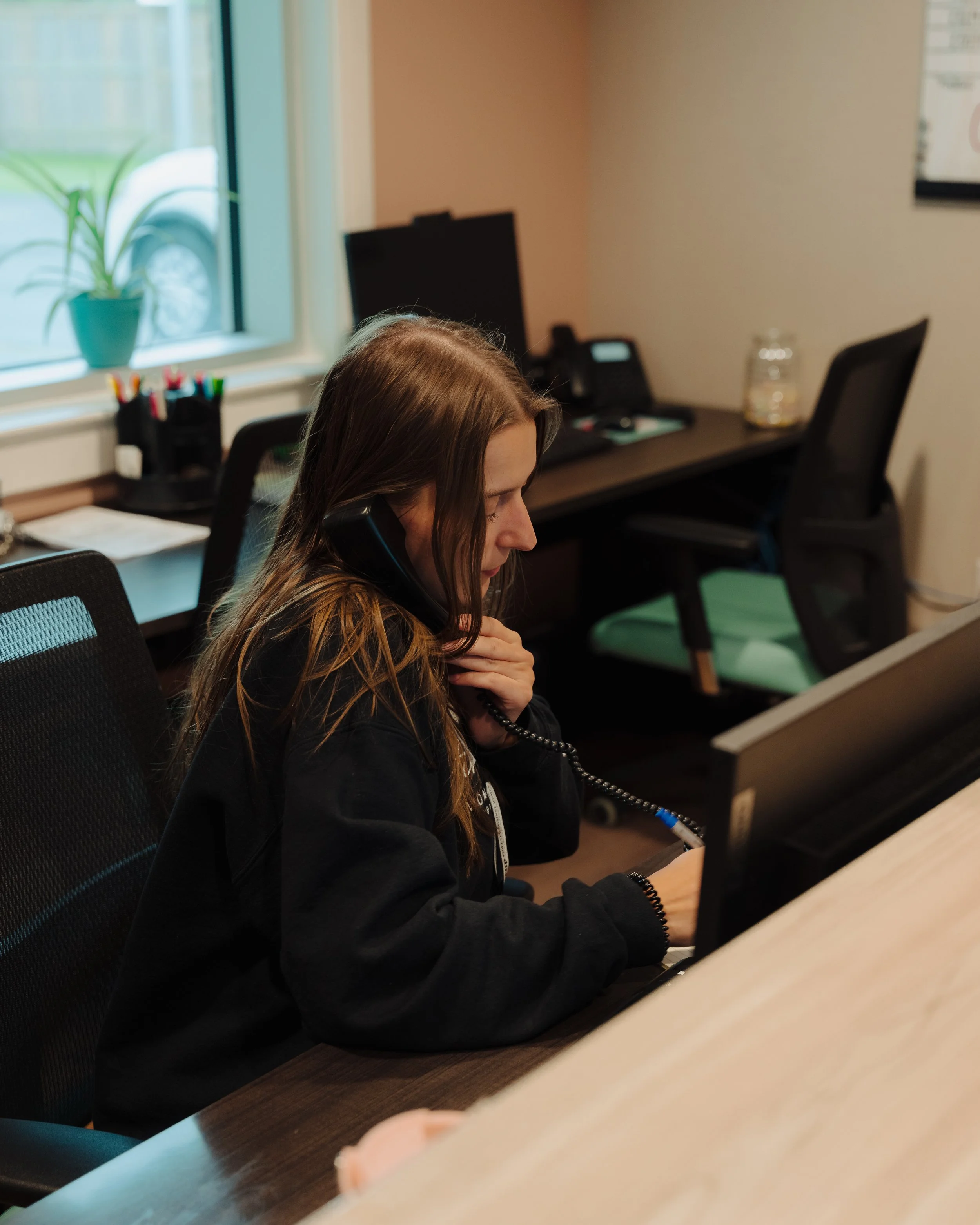 A young woman with long brown hair talking on a corded phone while looking at a computer monitor in an office.
