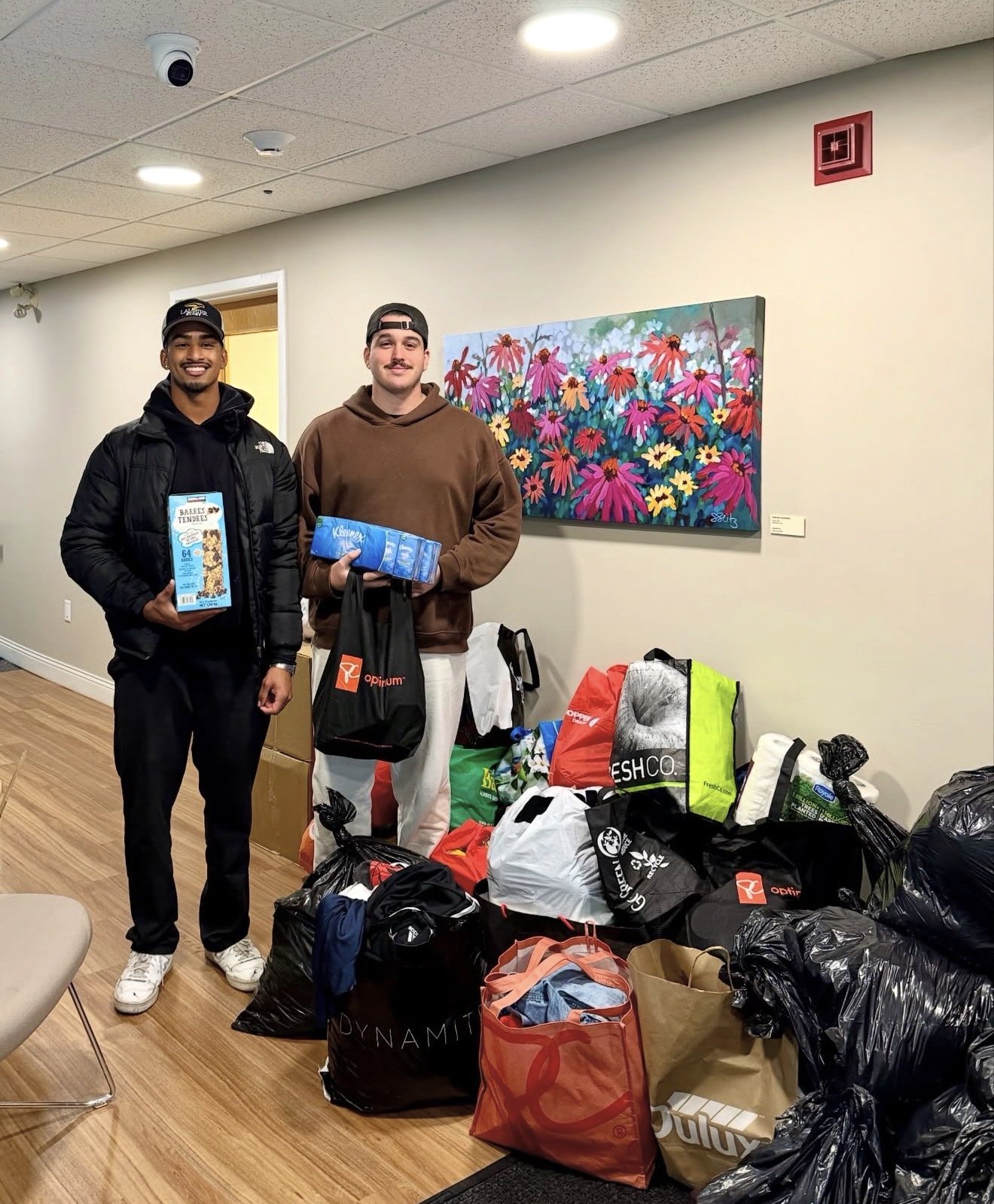 Two men stand in a room with bags of donated clothing and supplies, smiling and holding items, with a colorful floral painting on the wall behind them.