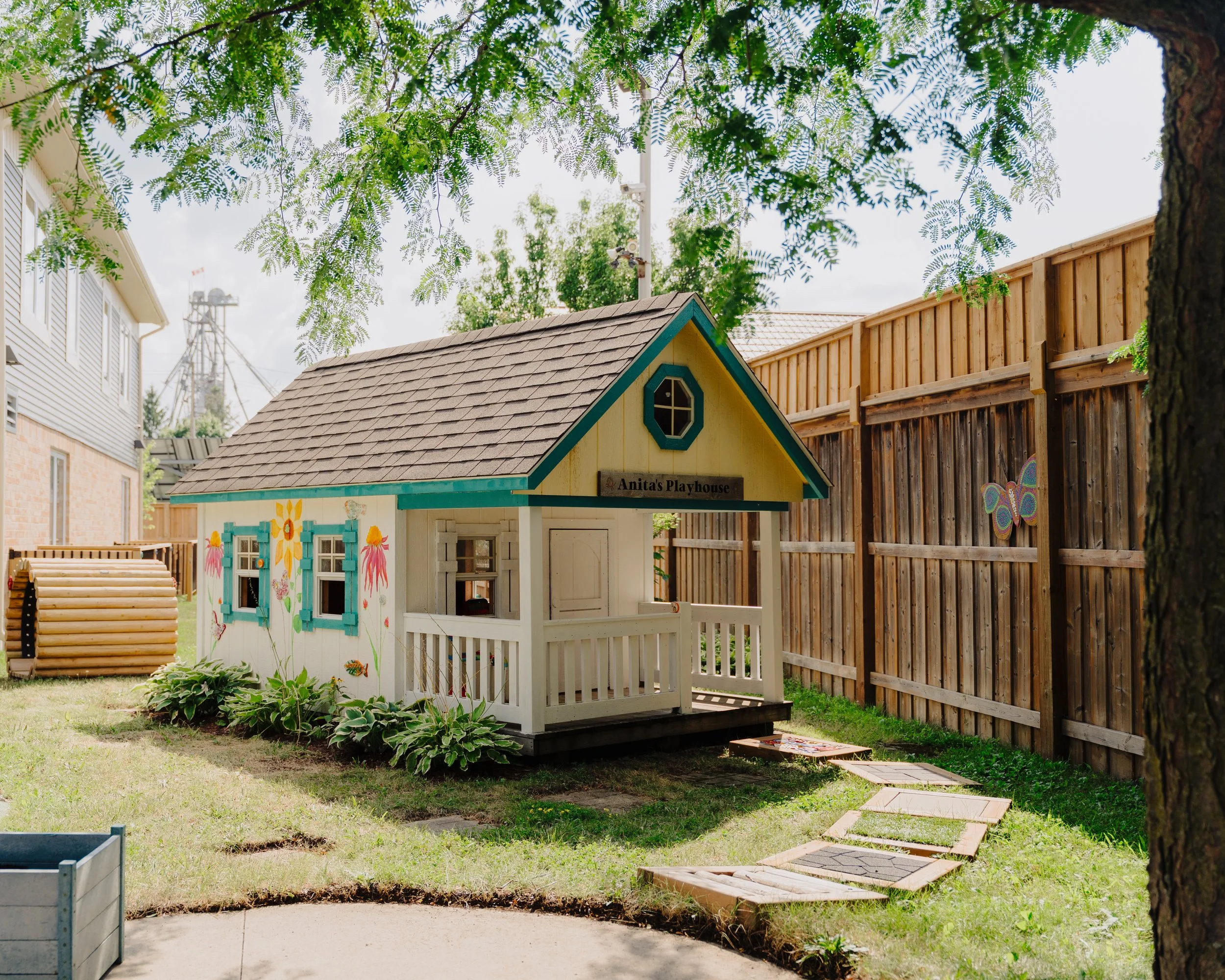 A small colorful playhouse labeled 'Anita's Playhouse' in a backyard, surrounded by a wooden fence, trees, and a garden path.