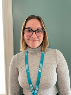 A woman with shoulder-length blonde hair, wearing glasses and a gray turtleneck sweater, smiling in front of a teal wall.